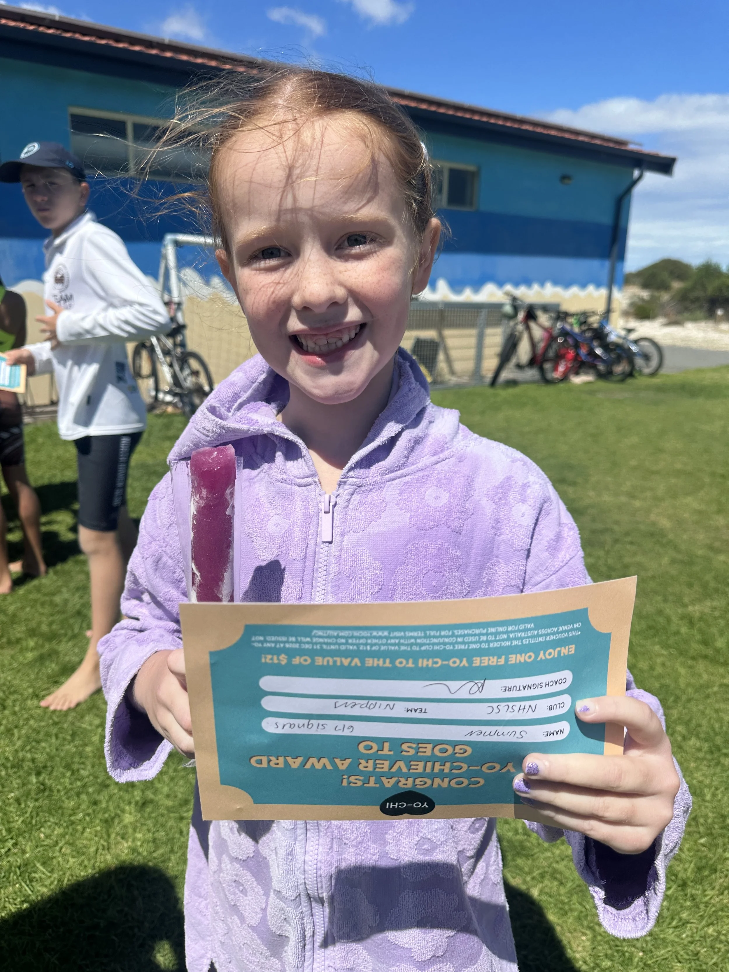 A young girl with red hair and a big smile holding an ice pop and a certificate outdoors on a sunny day, with a blue building and bicycles in the background.