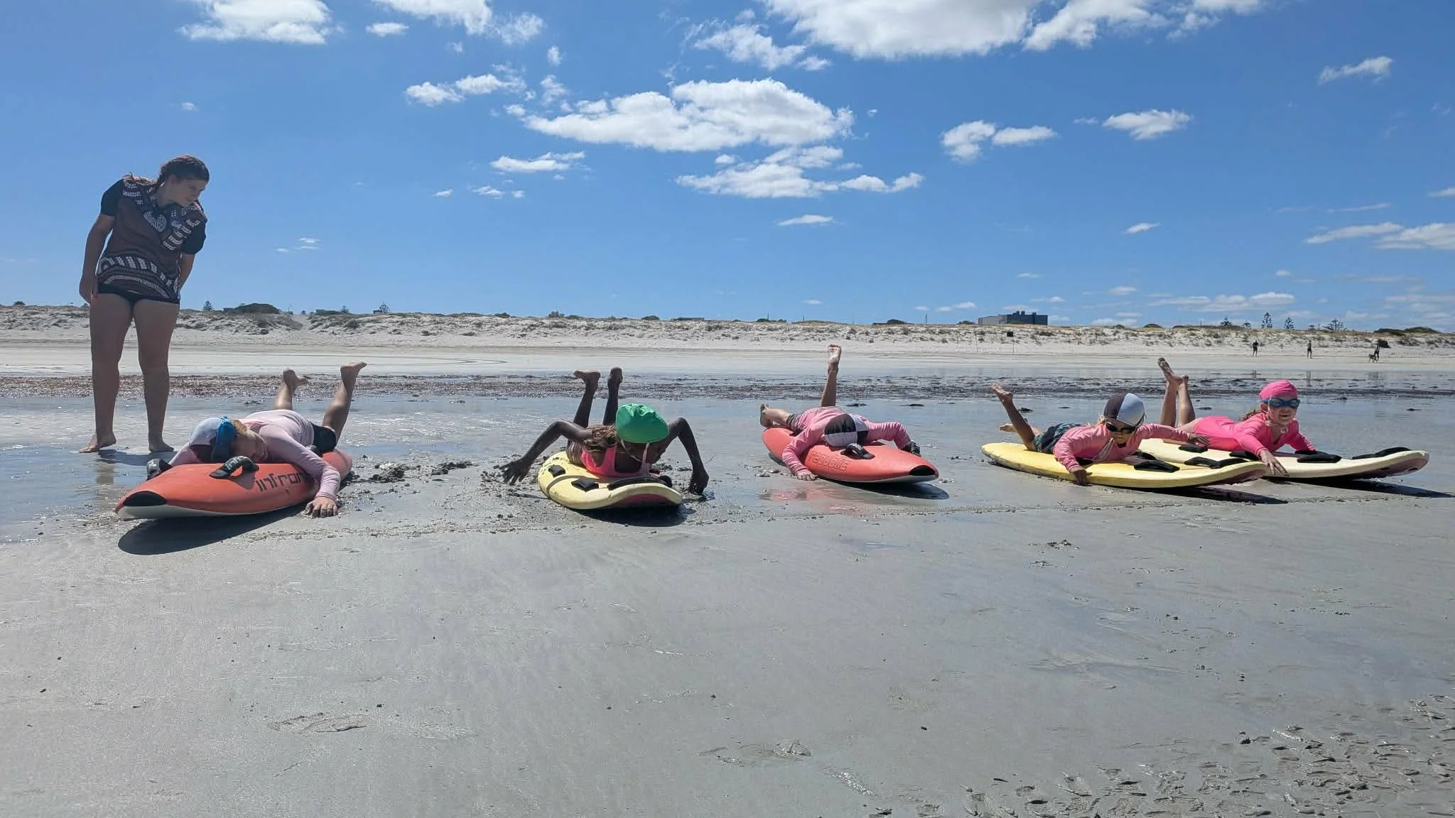Four children lying face down on surfboards on the sand at the beach, with an instructor standing nearby, under a partly cloudy blue sky.