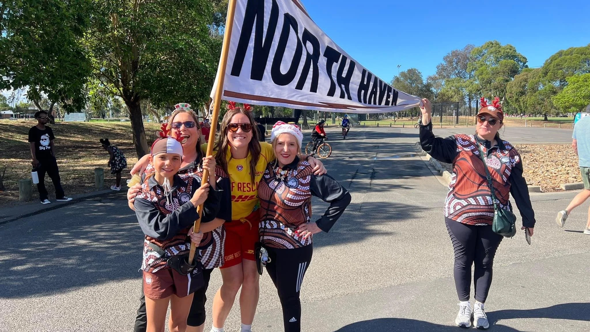 Group of women and children at a community event, holding a large banner that reads 'NORTHERN HAVEN,' with some wearing Christmas-themed accessories and some dressed in athletic wear, outdoors on a sunny day.