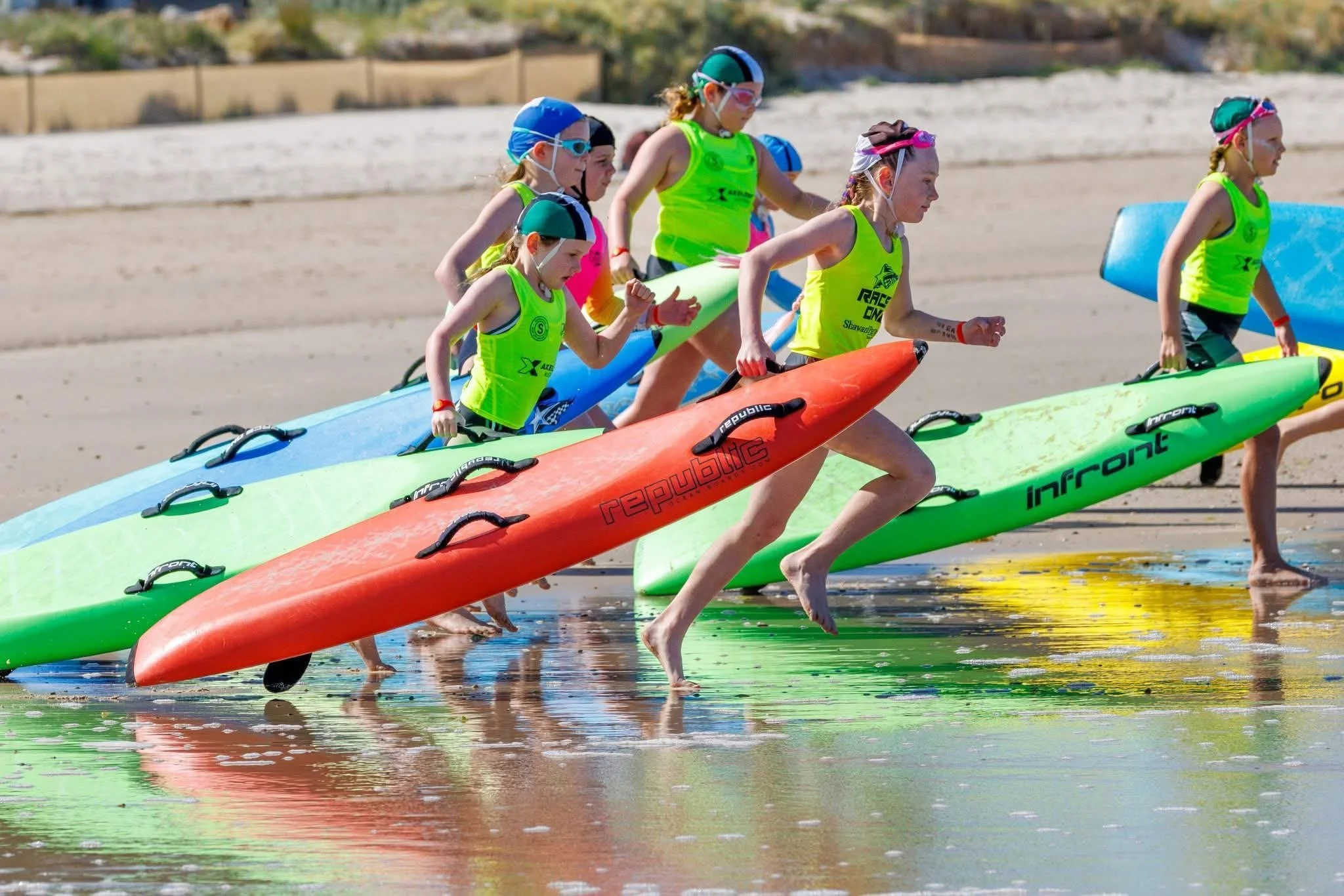 Young girls in bright green swimsuits and swimming caps running with colorful surfboards in shallow water at the beach during daytime.