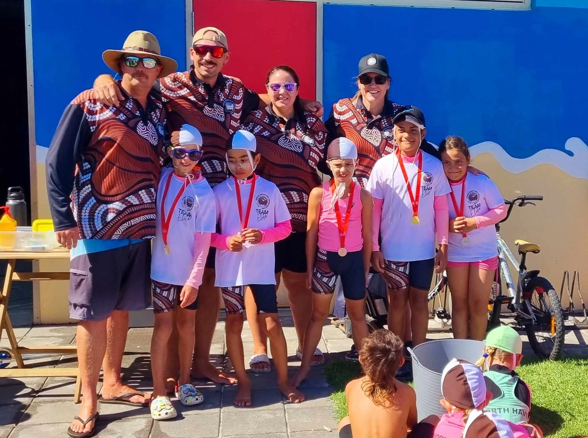 A group of adults and children posing outdoors after a sporting event, some children wearing medals, in front of a colorful wall, with a bicycle and a table nearby.