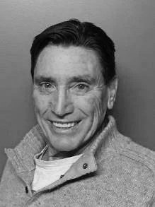 Black and white portrait of a smiling middle-aged man with dark hair, wearing a light-colored zip-up jacket.