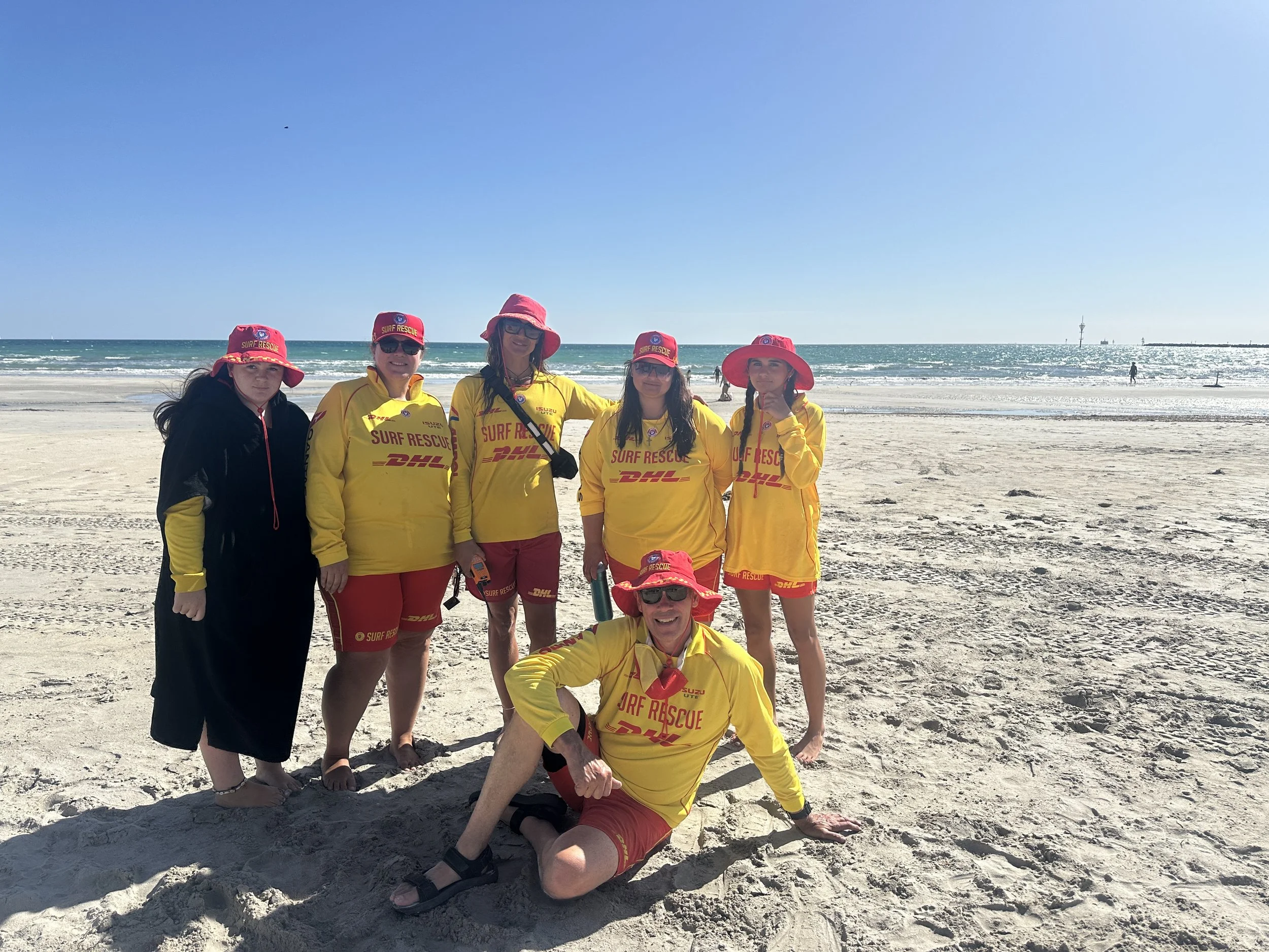 Group of six surf rescue team members on a beach, wearing yellow and red uniforms with DHL logos and red hats, with the ocean and clear sky in the background.