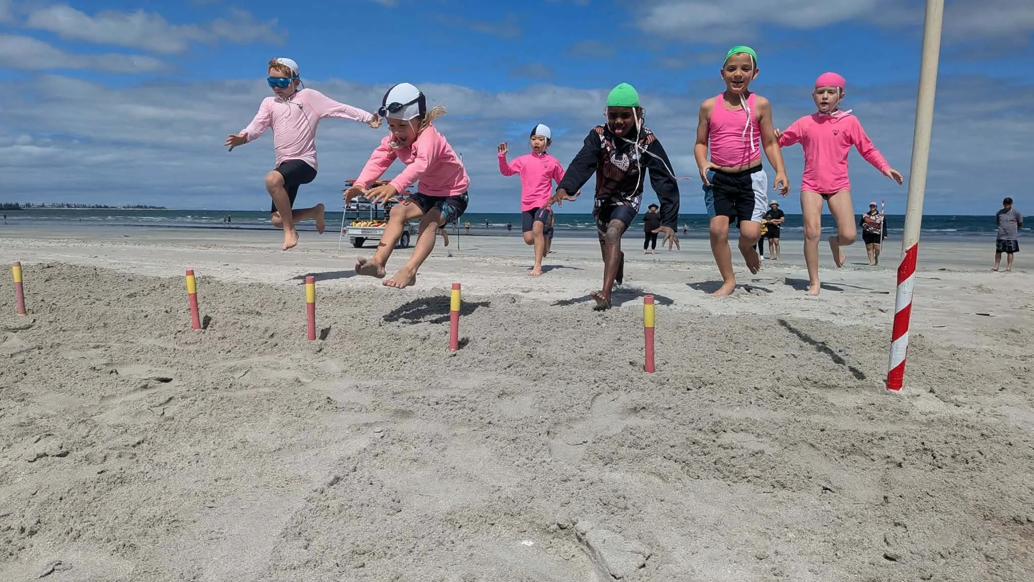 Children jumping over small hurdles on the beach under a blue sky.