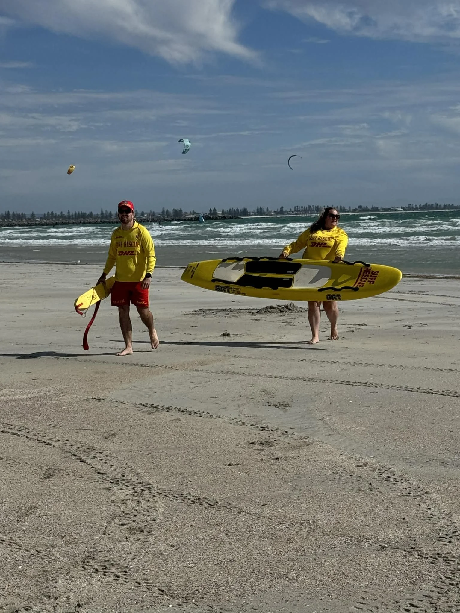 Two surf rescue personnel on a sandy beach, with one man holding a yellow rescue board and a woman holding a paddleboard. Several kite surfers are visible in the ocean under a partly cloudy sky.