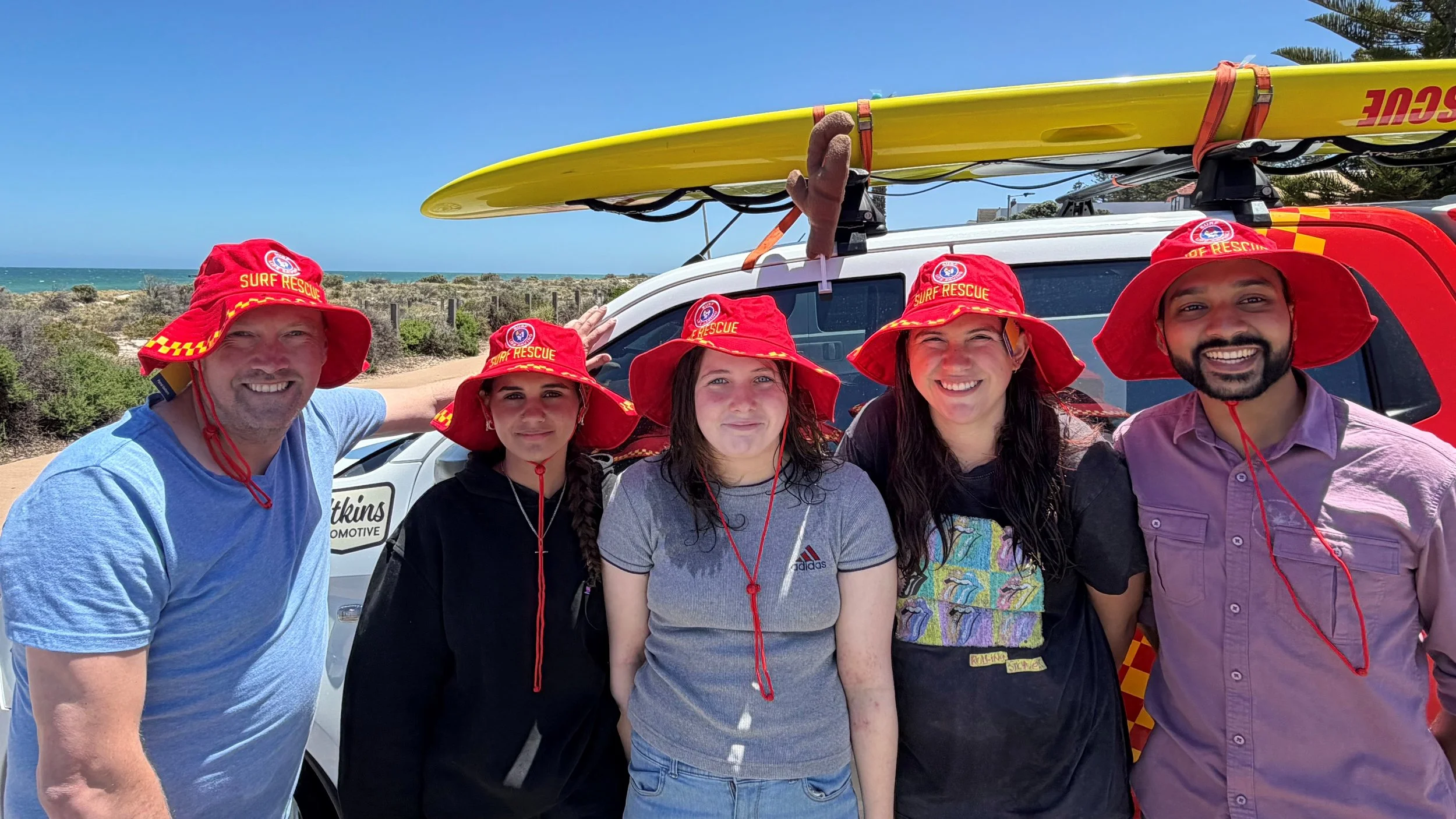A group of five people wearing red surf rescue hats smiling and standing in front of a rescue vehicle with surfboards on top, near the beach with a blue sky and ocean in the background.