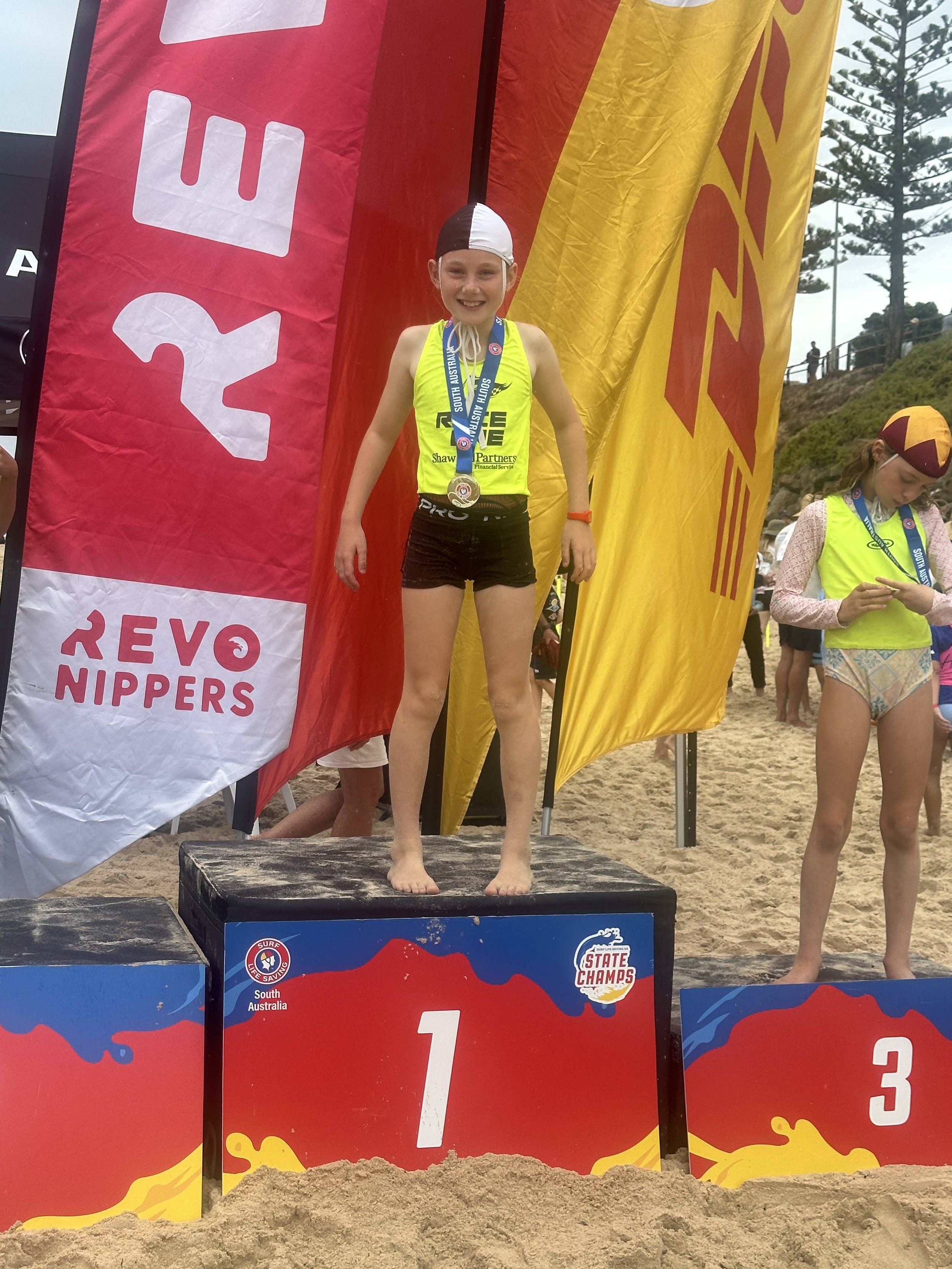 Young girl standing on a winner's podium at a beach surf competition, wearing a yellow sleeveless top, black shorts, a white and black swim cap, and a medal around her neck, smiling after winning first place.