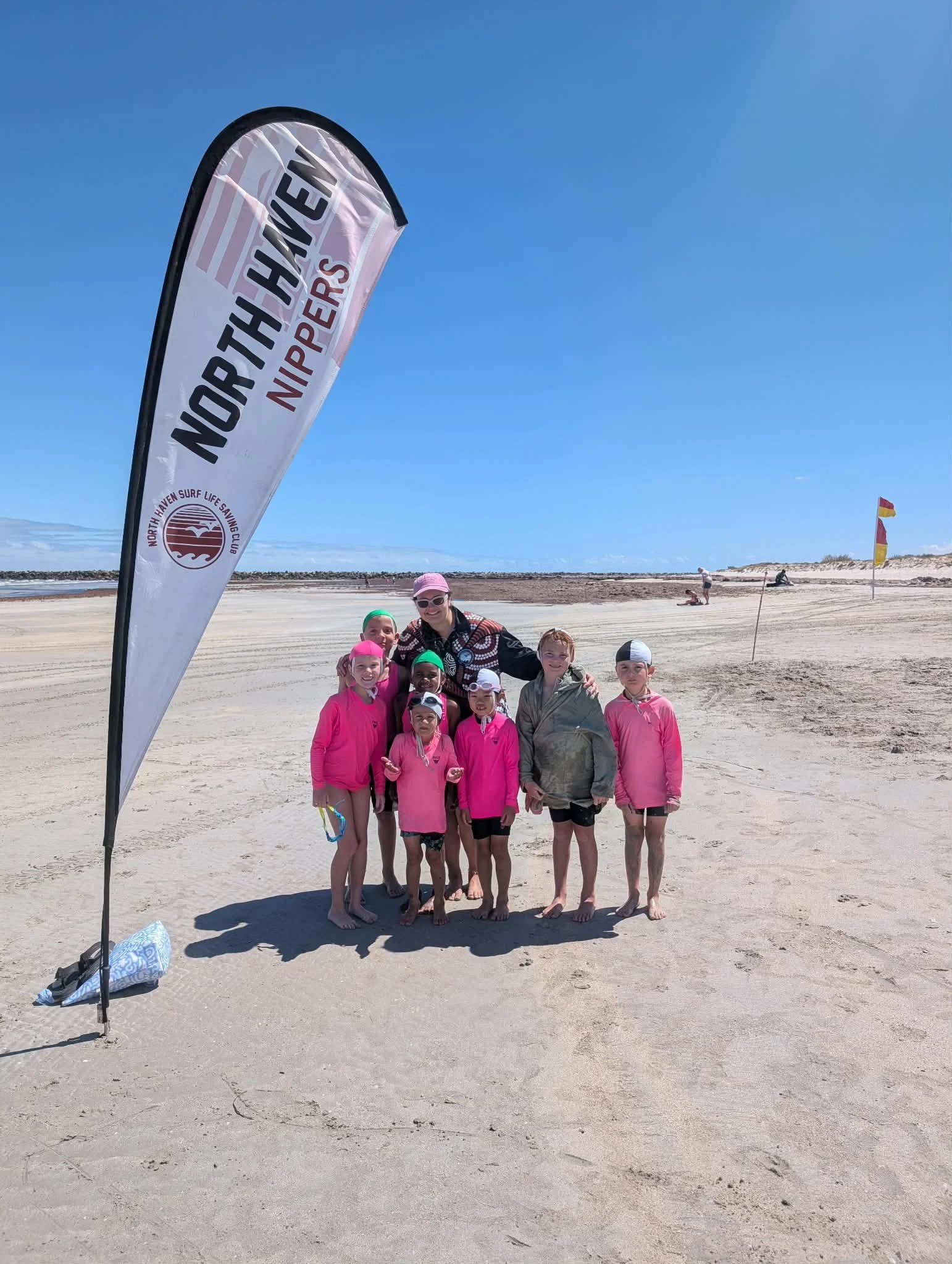 A group of children and an adult standing on the beach near a North Haven Surf Lifesaving Club flag, with a clear blue sky and some beach flags in the background.