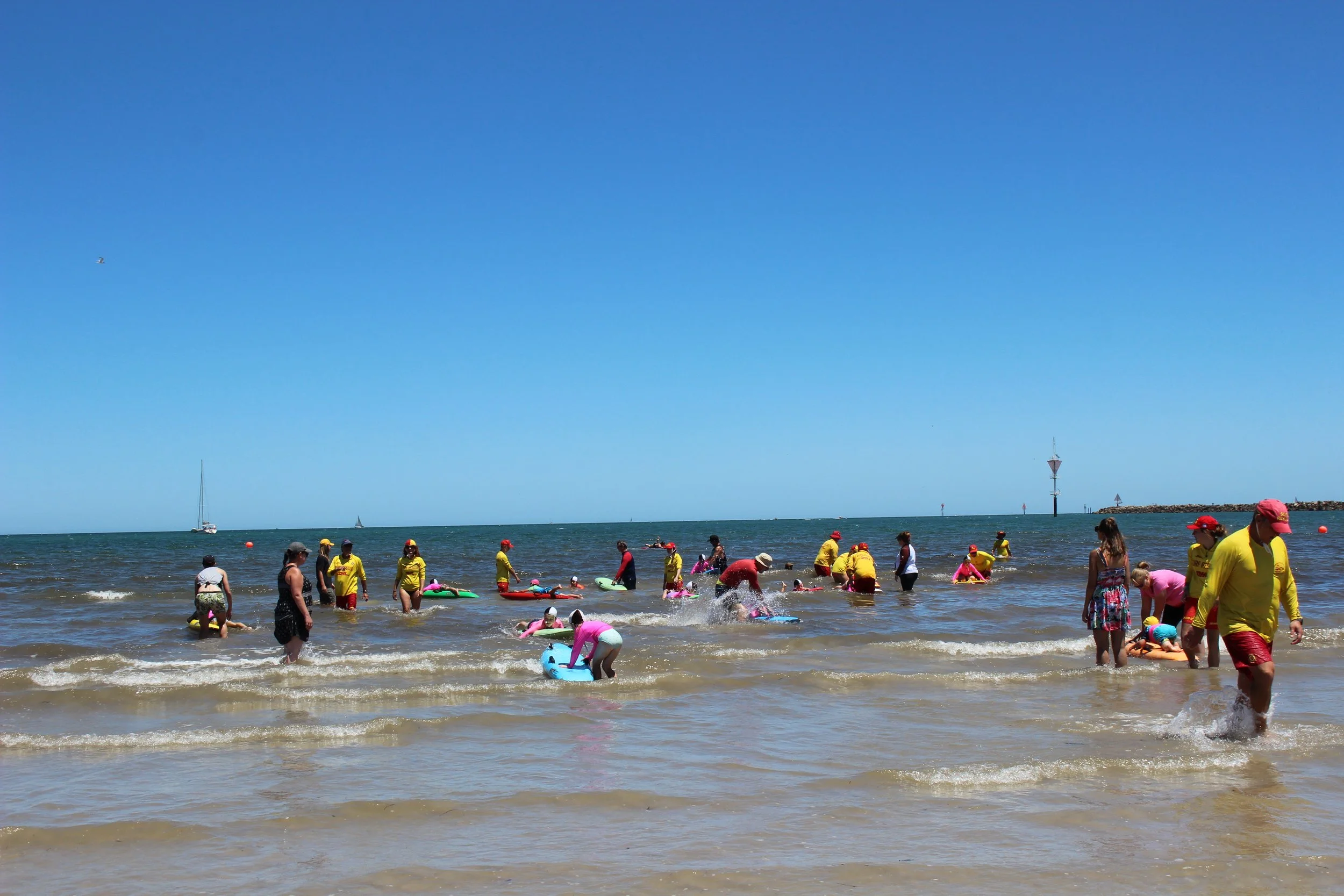 People enjoying a day at the beach, some with float toys, surrounded by clear blue water and sky, with a lighthouse and sailboats in the distance.