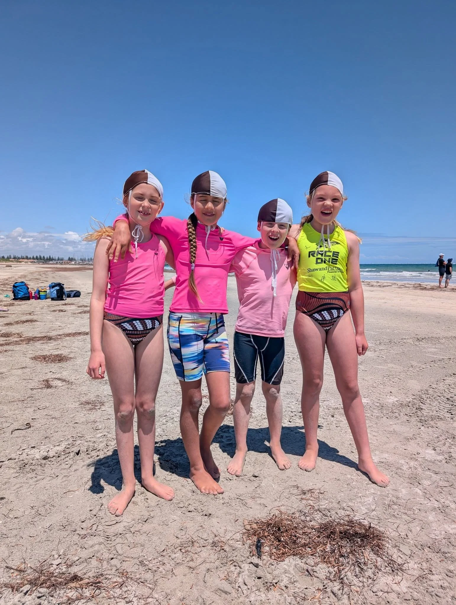 Four young girls standing arm-in-arm on a beach, wearing swimsuits and swim caps, smiling at the camera under a clear blue sky with a few scattered clouds. Beach gear is visible behind them, and the ocean is in the background.