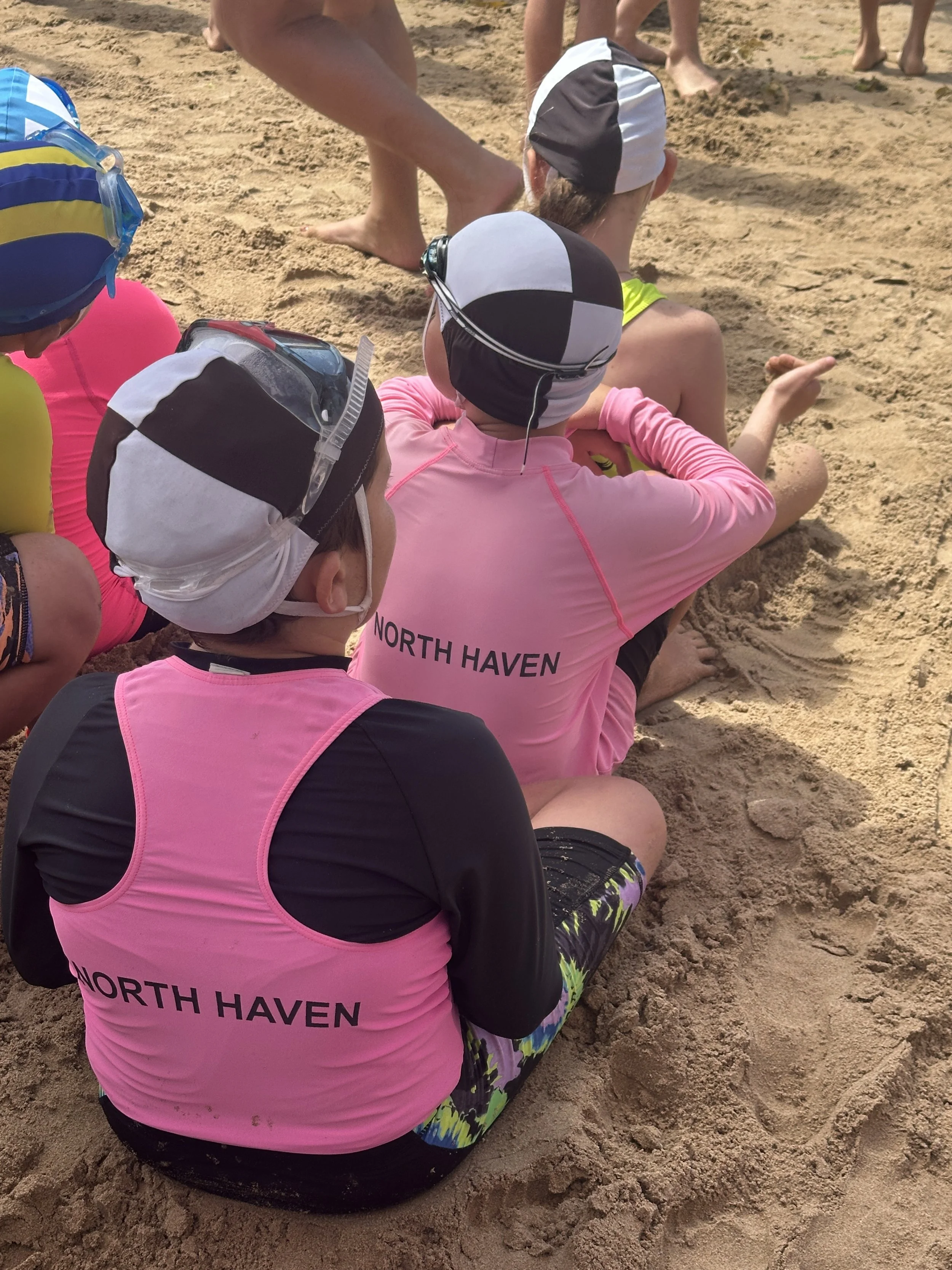Group of children sitting on sandy beach, wearing pink and black swimwear with 'North Haven' printed on the back, and swim caps with goggles, likely listening to instructions or a story.