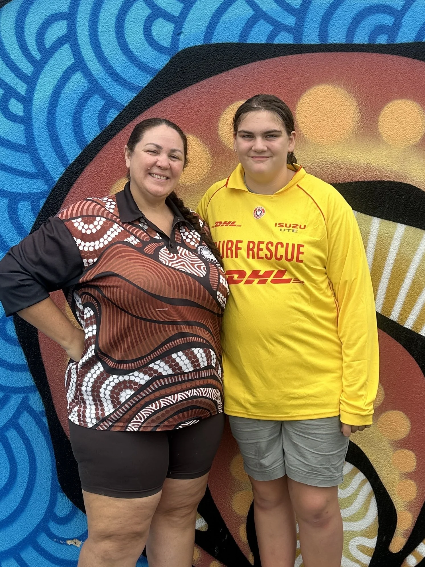 Two women standing together in front of a colorful mural. The woman on the left is wearing a patterned shirt and black shorts, smiling with her hand on her hip. The woman on the right is wearing a yellow surf rescue shirt and gray shorts, with a neutral expression.