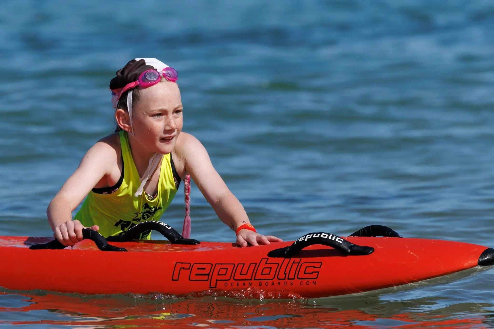 A young girl in a yellow tank top paddling a red paddleboard on water, wearing pink goggles and a white hat.