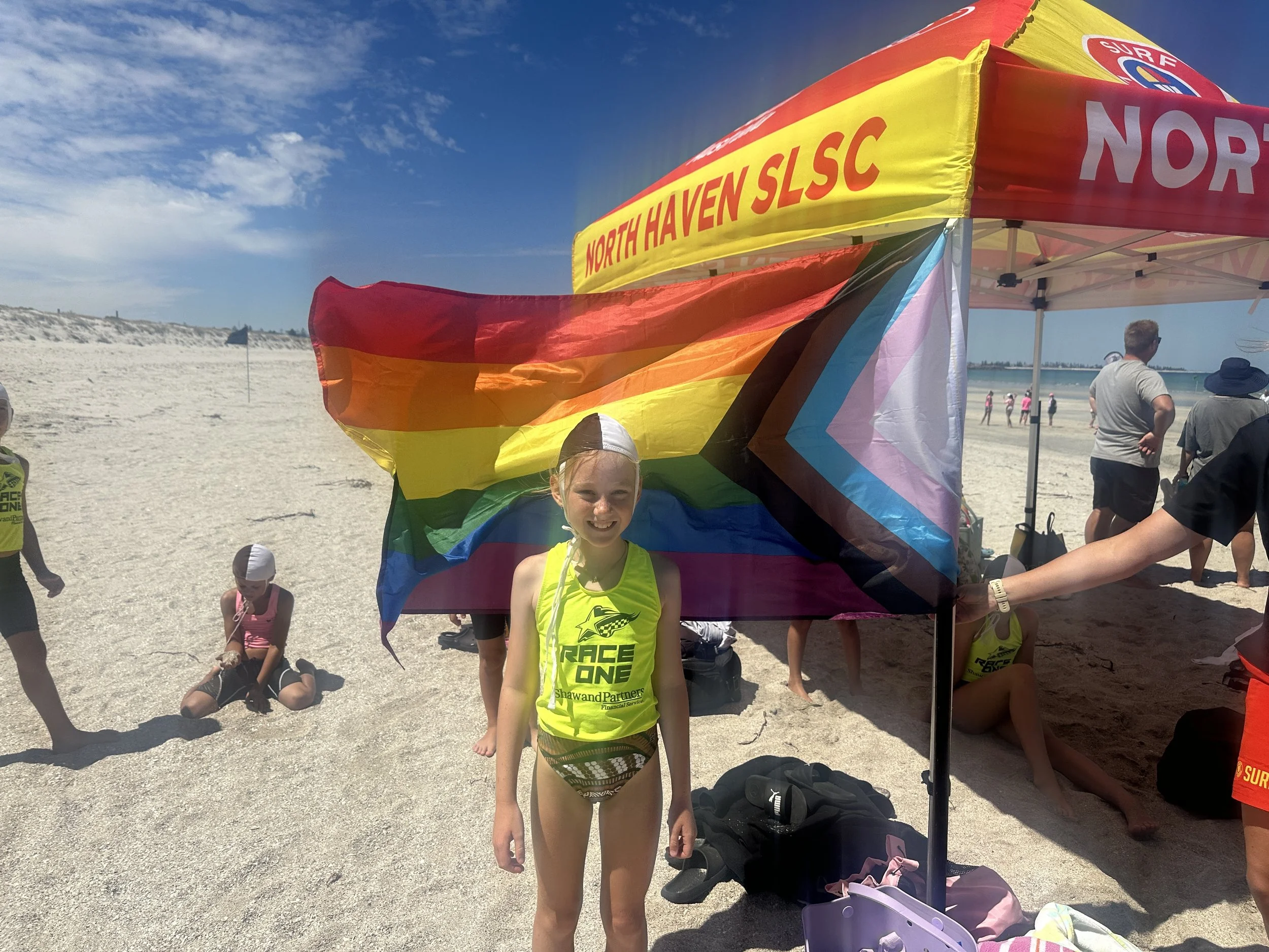 Young girl smiling at the camera, standing next to a large rainbow flag and a yellow tent with 'North Haven SLSC' written on it, at the beach on a sunny day.