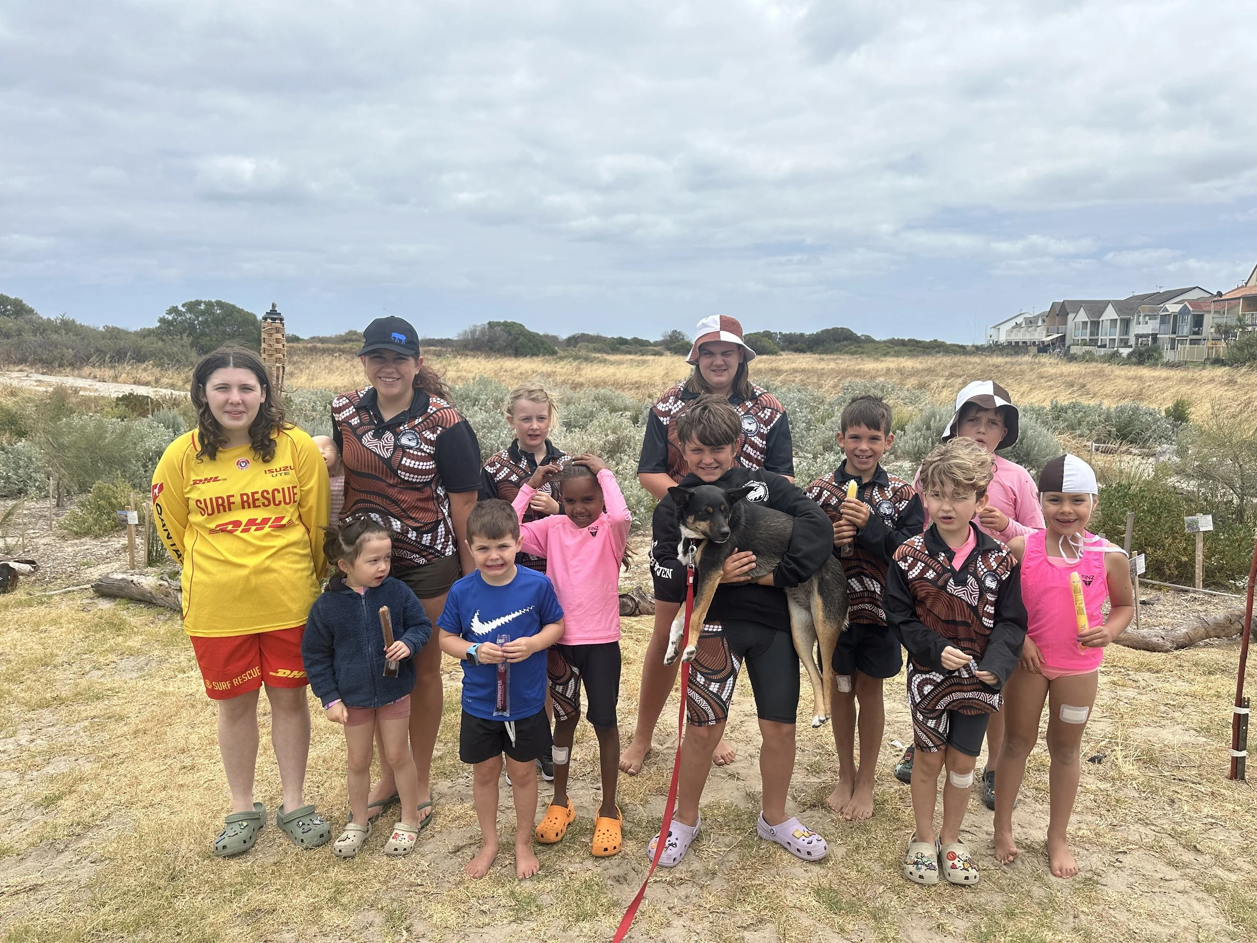 Group of children and young adults outdoors, some in uniform and others in casual clothing, holding dog treats and a dog, in a natural landscape with grass and shrubs, overcast sky.