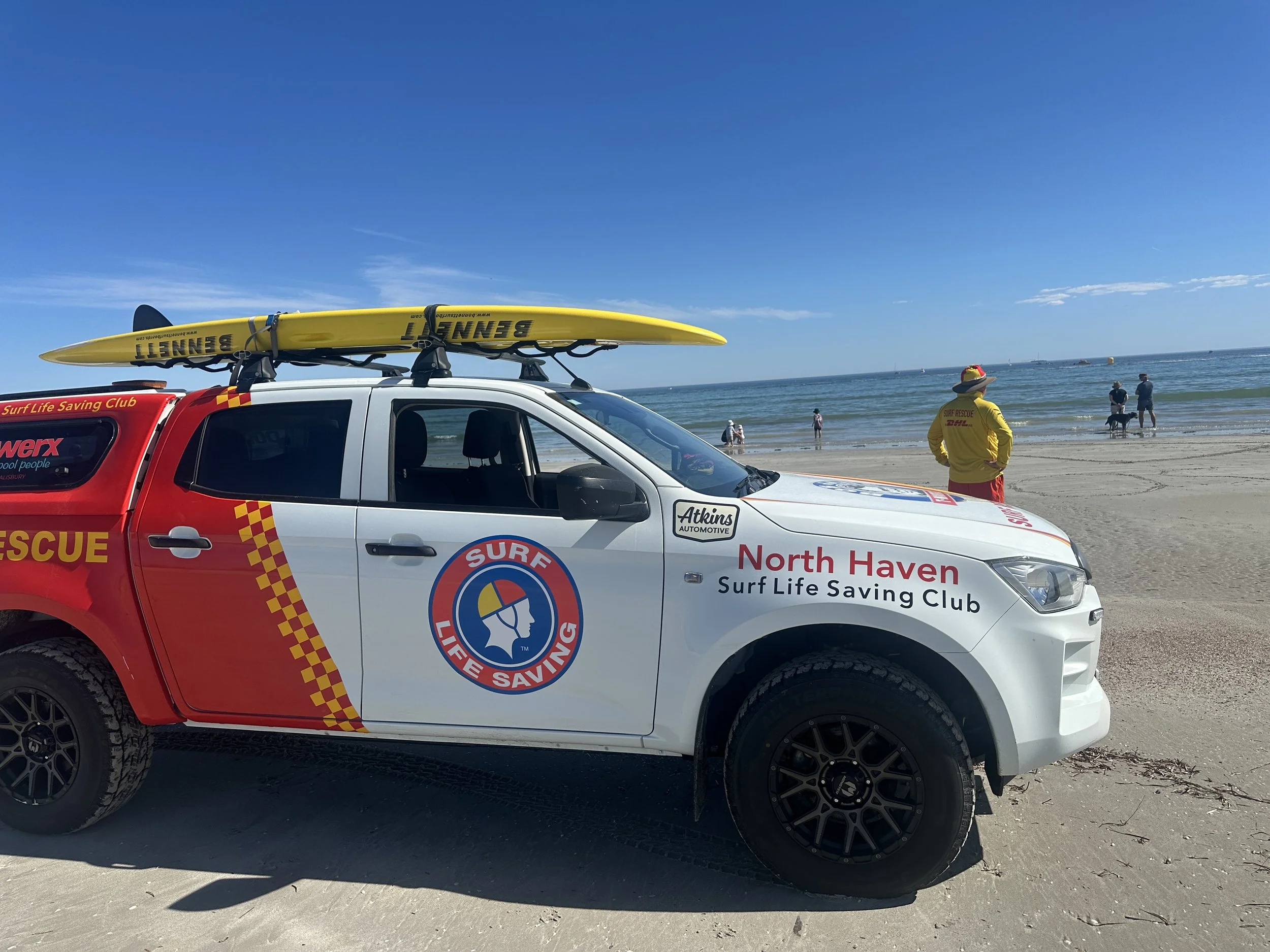 A North Haven Surf Life Saving Club rescue vehicle parked on the sandy beach near the ocean, with a yellow surfboard on top. A rescue worker in yellow and red stands near the water, and several people are on the beach near the shoreline.