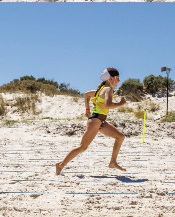 A woman in a yellow sports top and black shorts running on a sandy beach during a race, with a blue sky and dunes in the background.