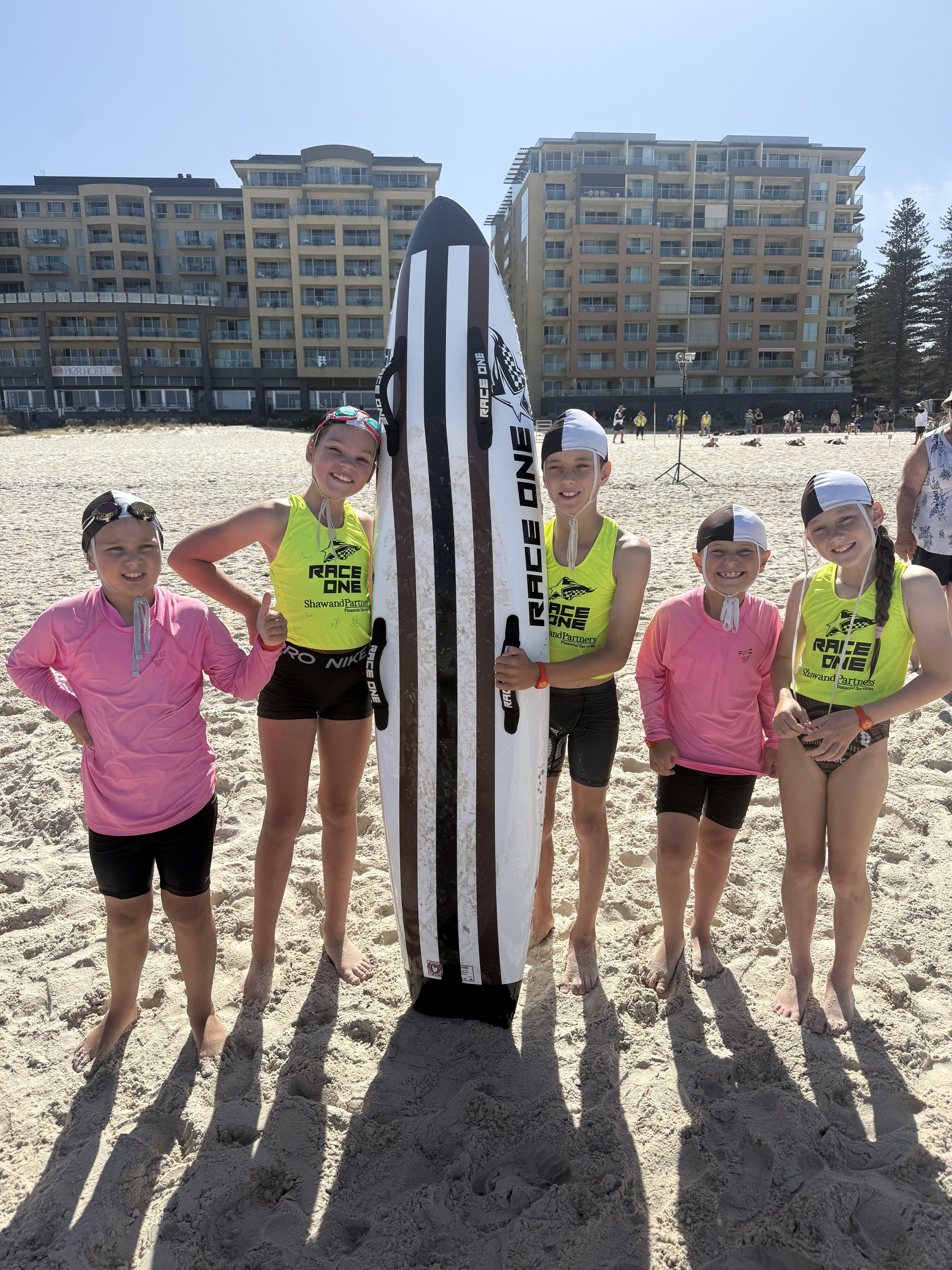 Four young girls in swimwear and neon shirts on a sandy beach, smiling and posing with a large surfboard.