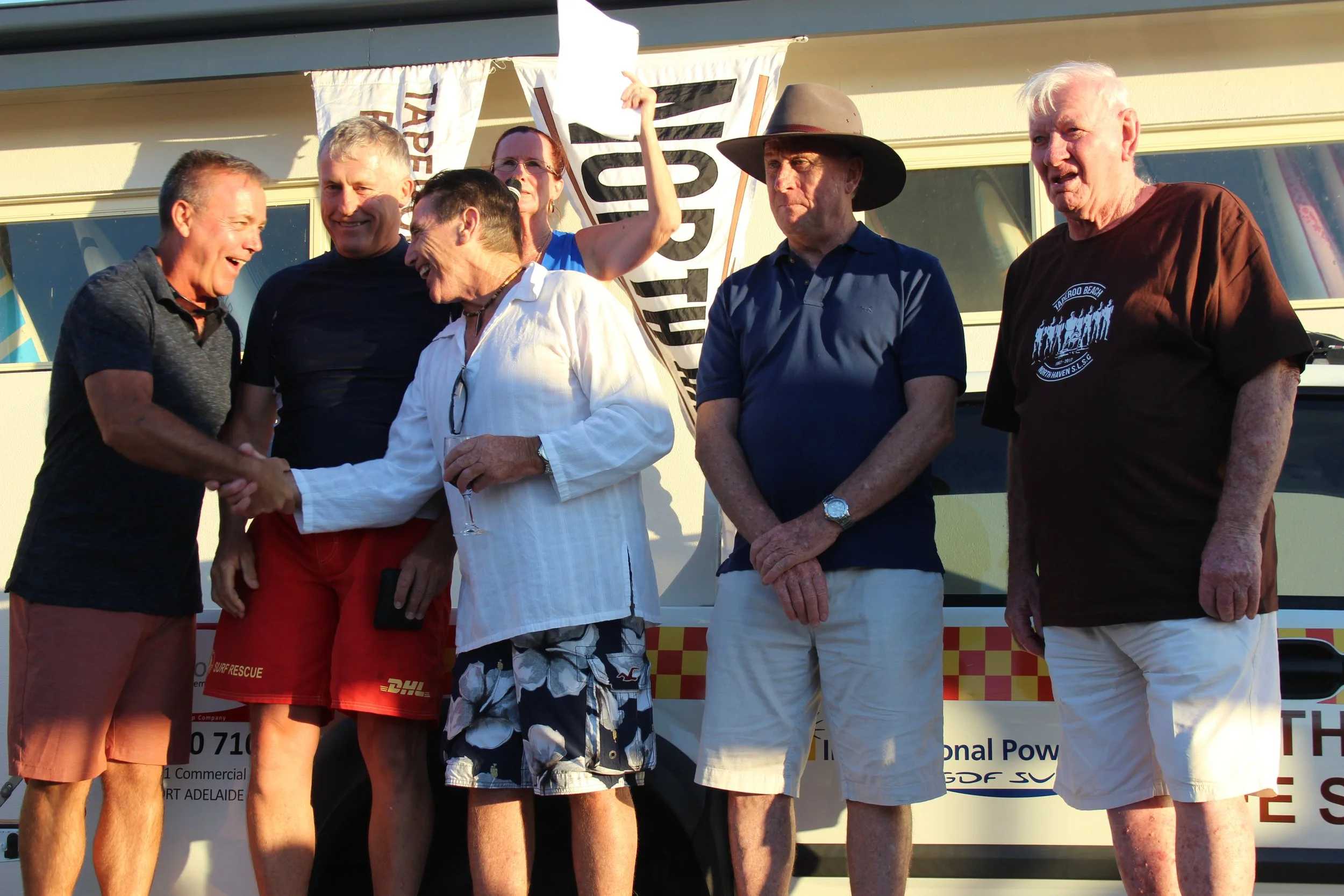 Group of six men standing in front of a bus, two of them shaking hands and smiling, during an outdoor event.