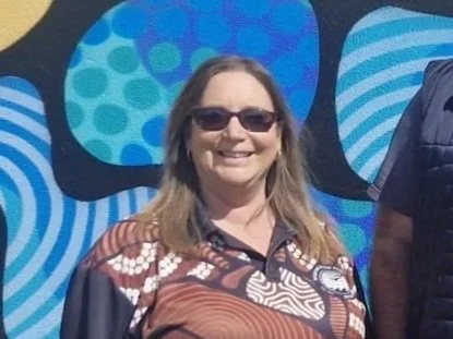 Woman with long hair wearing sunglasses and a patterned shirt, smiling outdoors with colorful abstract art in the background.