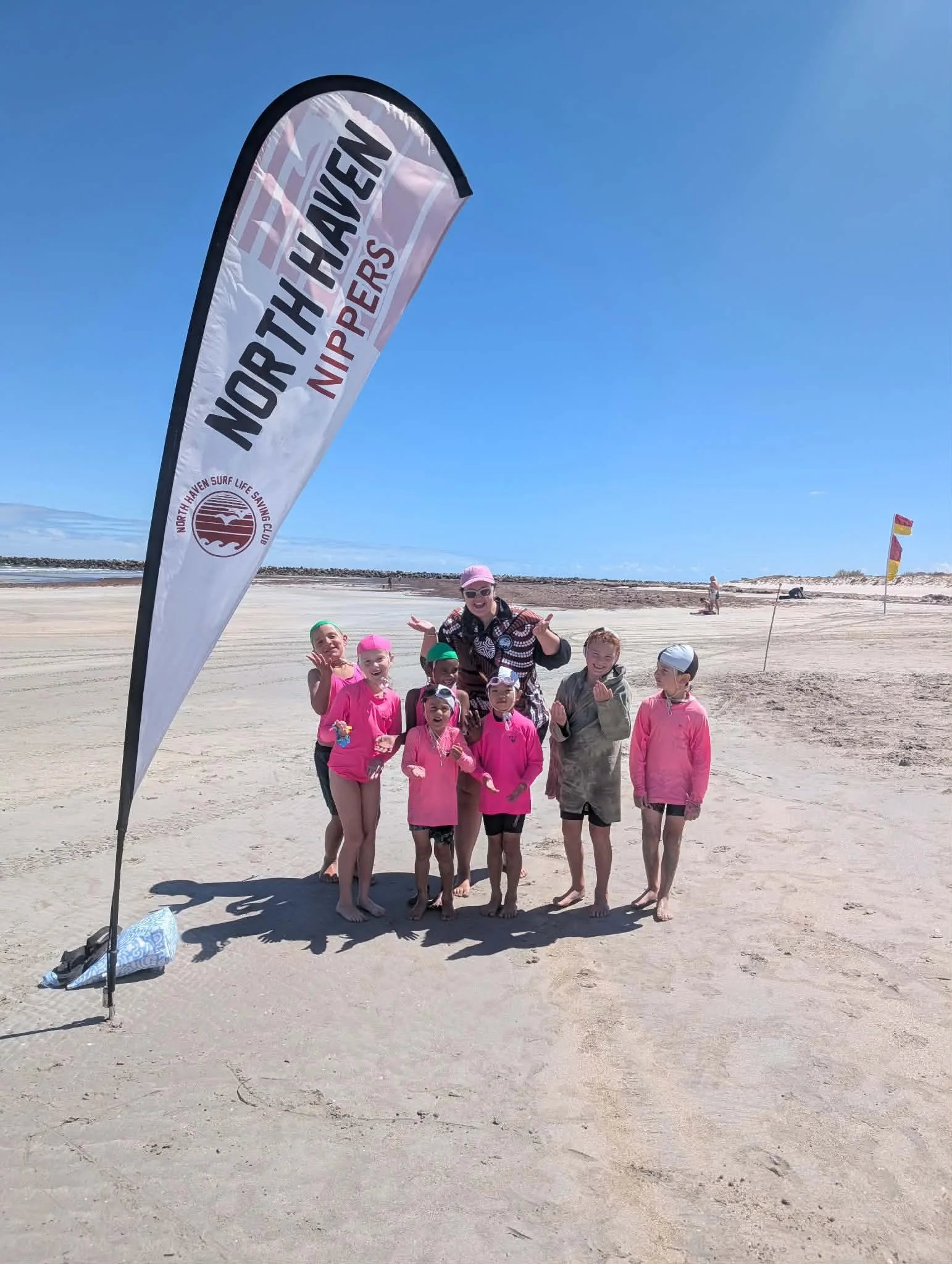 Group of children and a woman on a beach, holding medals, near a North Haven Nippers flag from North Haven Surf Life Saving Club. The sky is clear and blue.