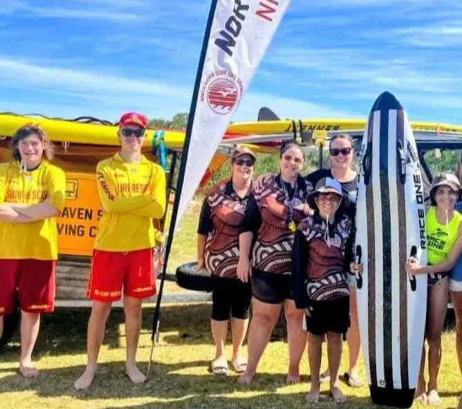 Group of people at a summer surf festival, some wearing yellow and red surf team uniforms, other in patterned shirts, with surfboards and a boat in the background.