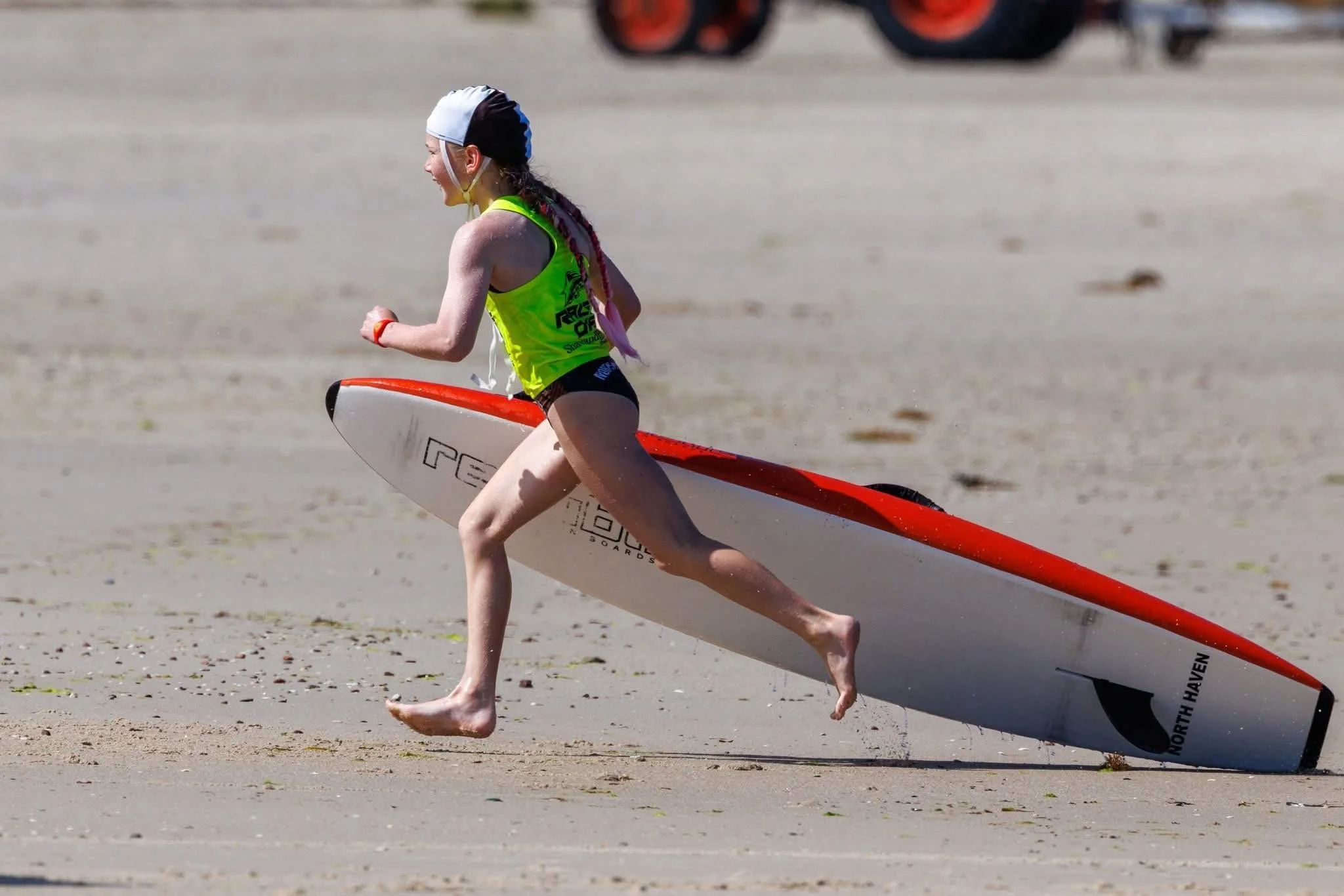A young girl in a yellow vest and bathing cap runs barefoot on a sandy beach while carrying a stand-up paddleboard.