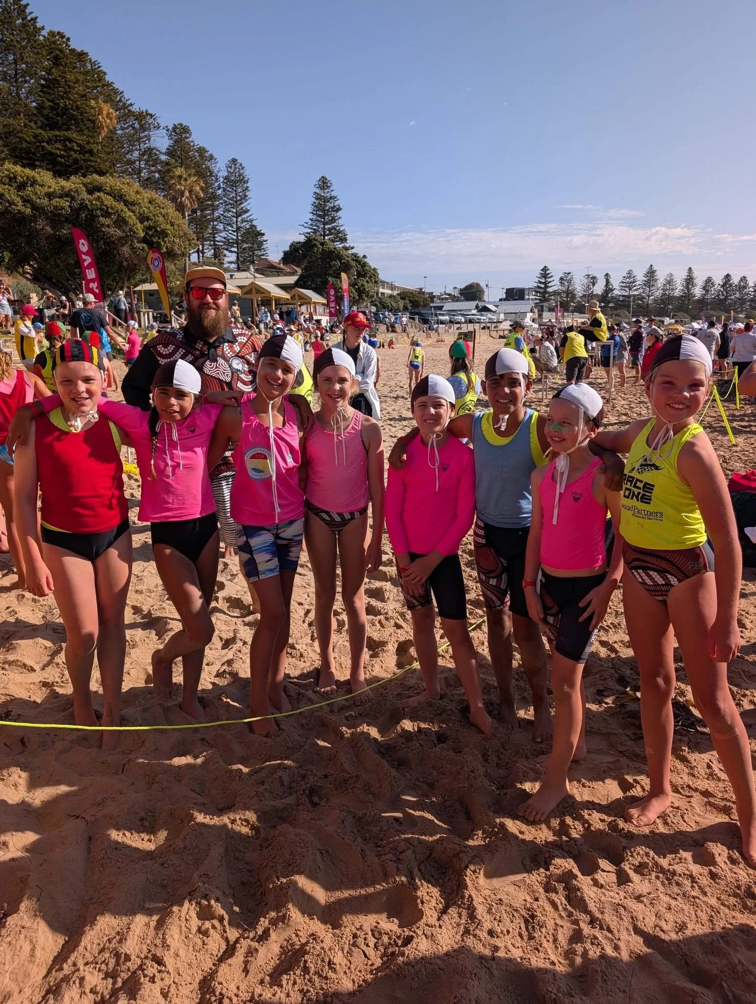 A group of young children in colorful swimsuits and swim caps standing together on a sandy beach with a man, likely their coach or instructor, against a background of beachgoers, tents, flags, and trees under a clear blue sky.