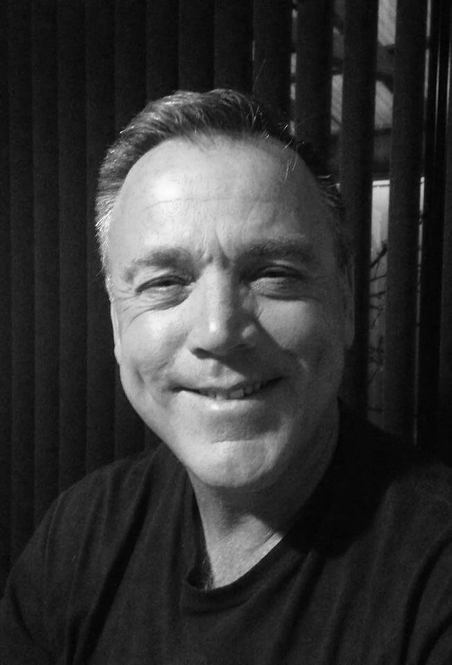 Black and white photo of a smiling man with short hair, wearing a dark shirt, sitting in front of a wooden background.