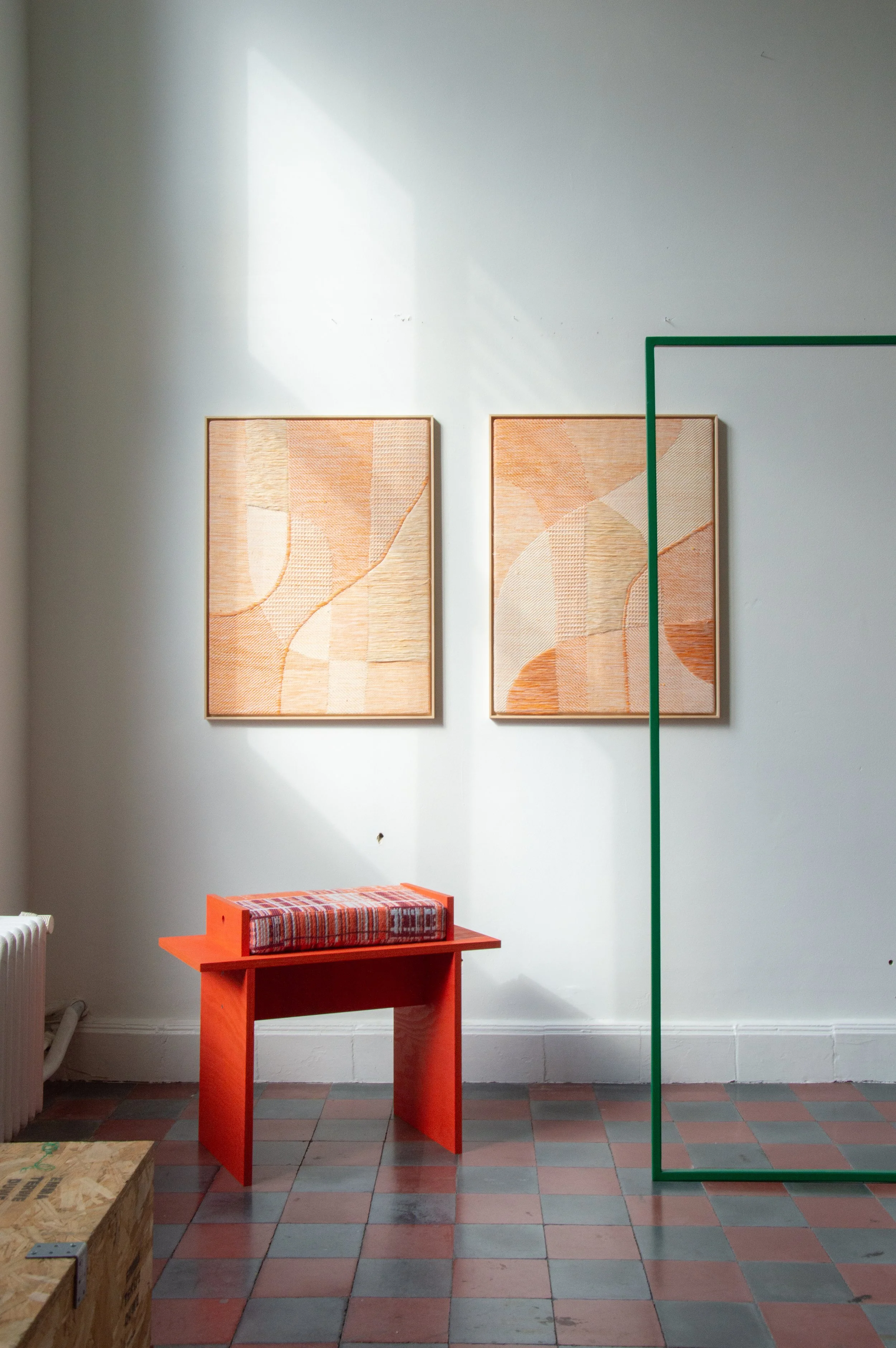 Twee abstracte kunstwerken hangen aan een witte muur, een kleine rode tafel met een gestreepte kussen eronder staat voor de muur, en een groene frame staat rechts in de afbeelding.