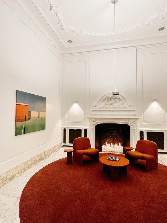 Een elegante witte kamer met een open haard, twee oranje stoelen, een ronde salontafel en een rode halfronde vloerkleed. Er is een schilderij aan de muur en zachte verlichting.