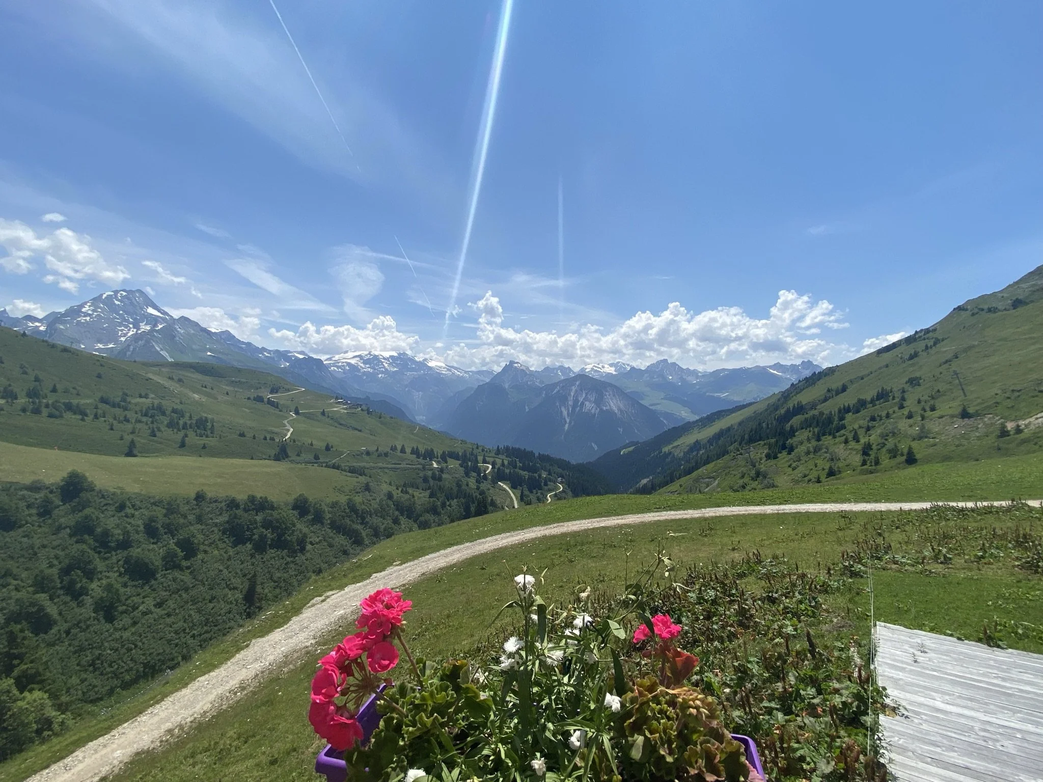 Paysage de montagne avec des sommets enneigés, des collines verdoyantes, un ciel bleu clair avec quelques nuages et plusieurs voies de chemin de fer traversant la vallée