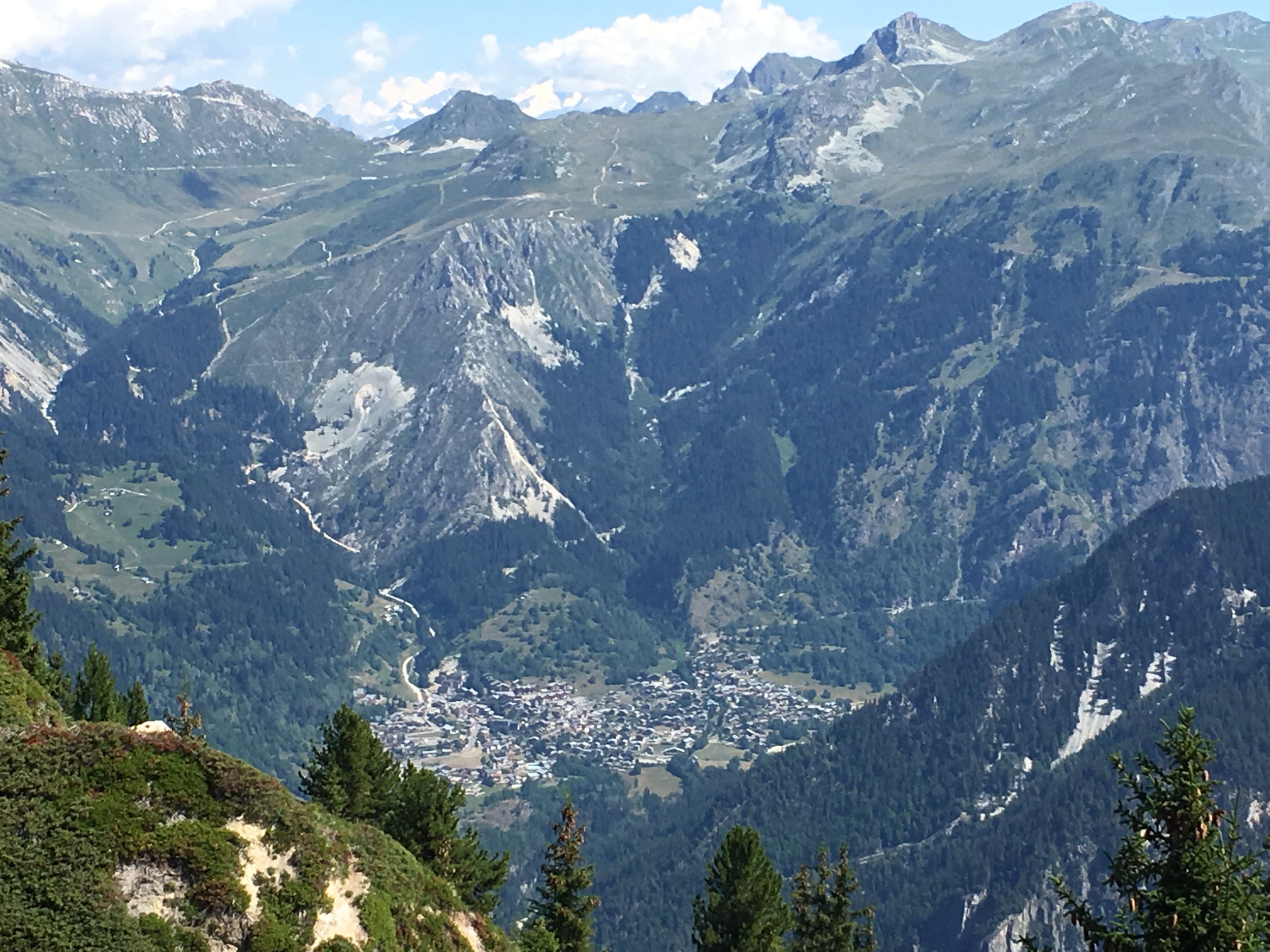 Paysage de montagnes avec village en vallée, arbres verts au premier plan, sommet rocheux avec quelques traces de neige.