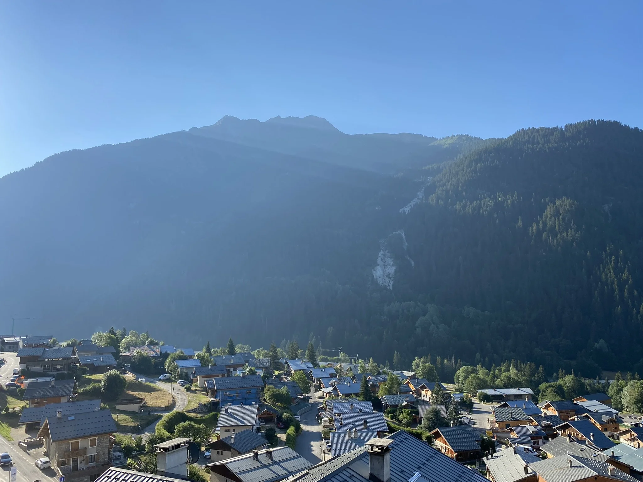 Vue panoramique d'un village alpin avec des maisons en bois et en pierre, entouré de montagnes dans un paysage ensoleillé.