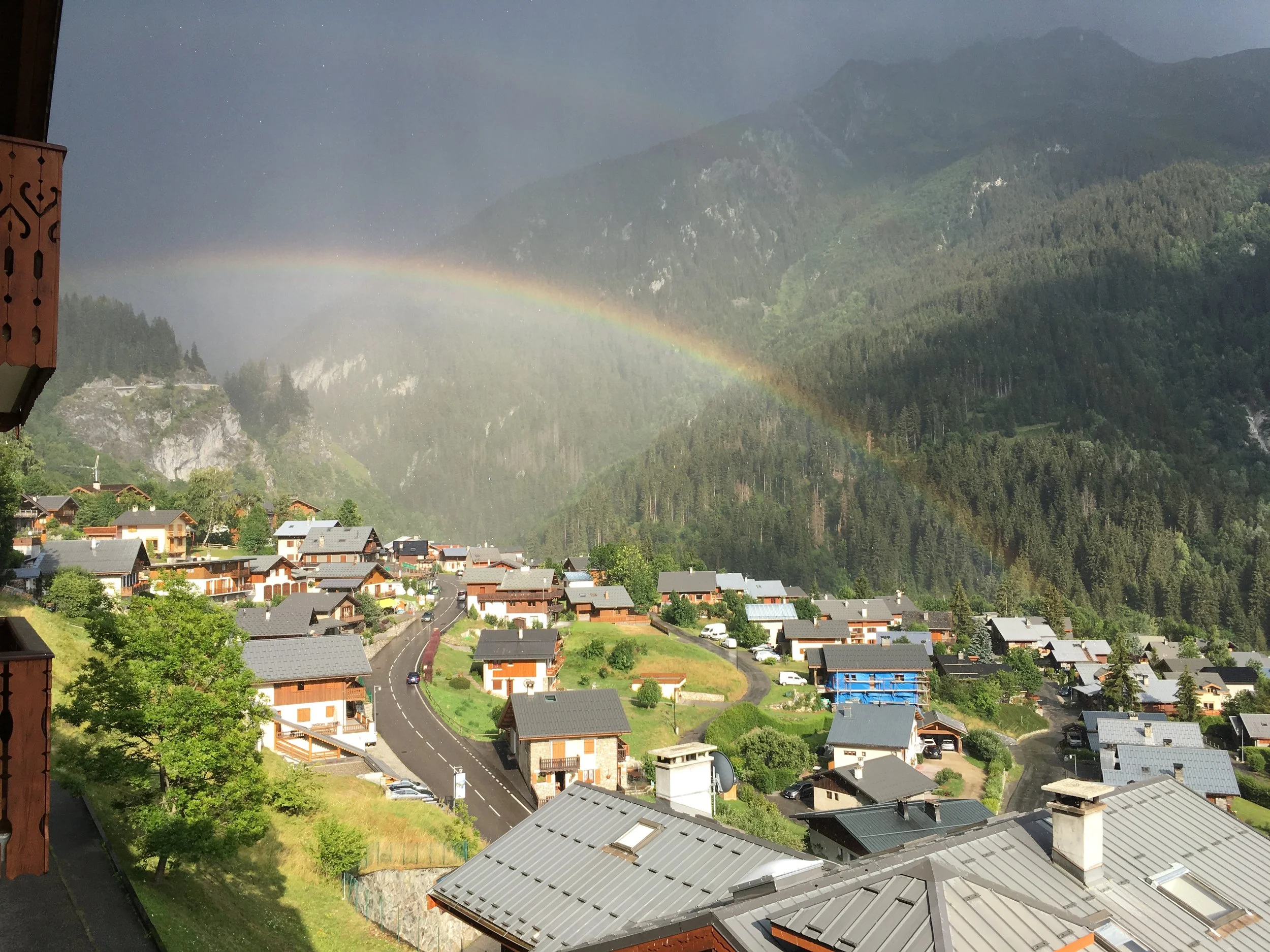 Village montagnard avec maisons en bois, routes sinueuses, forêt dense et arc-en-ciel dans le ciel.
