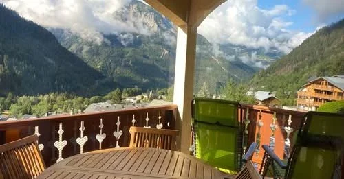 Balcon avec une table en bois et des chaises, vue sur des montagnes et un village alpin sous un ciel partiellement nuageux.