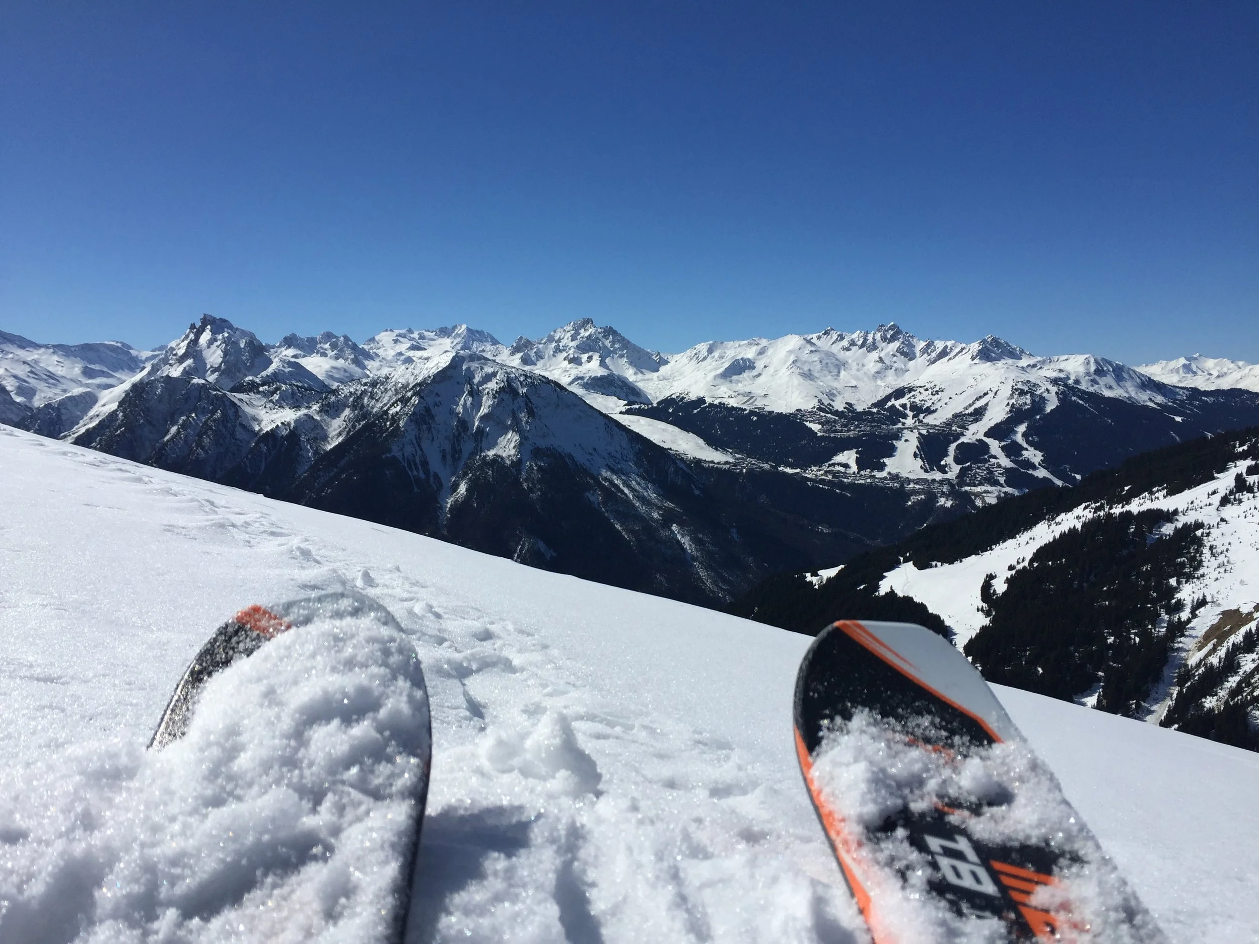 Vue de skis sur une pente enneigée avec une chaîne de montagnes enneigées en arrière-plan sous un ciel bleu clair.
