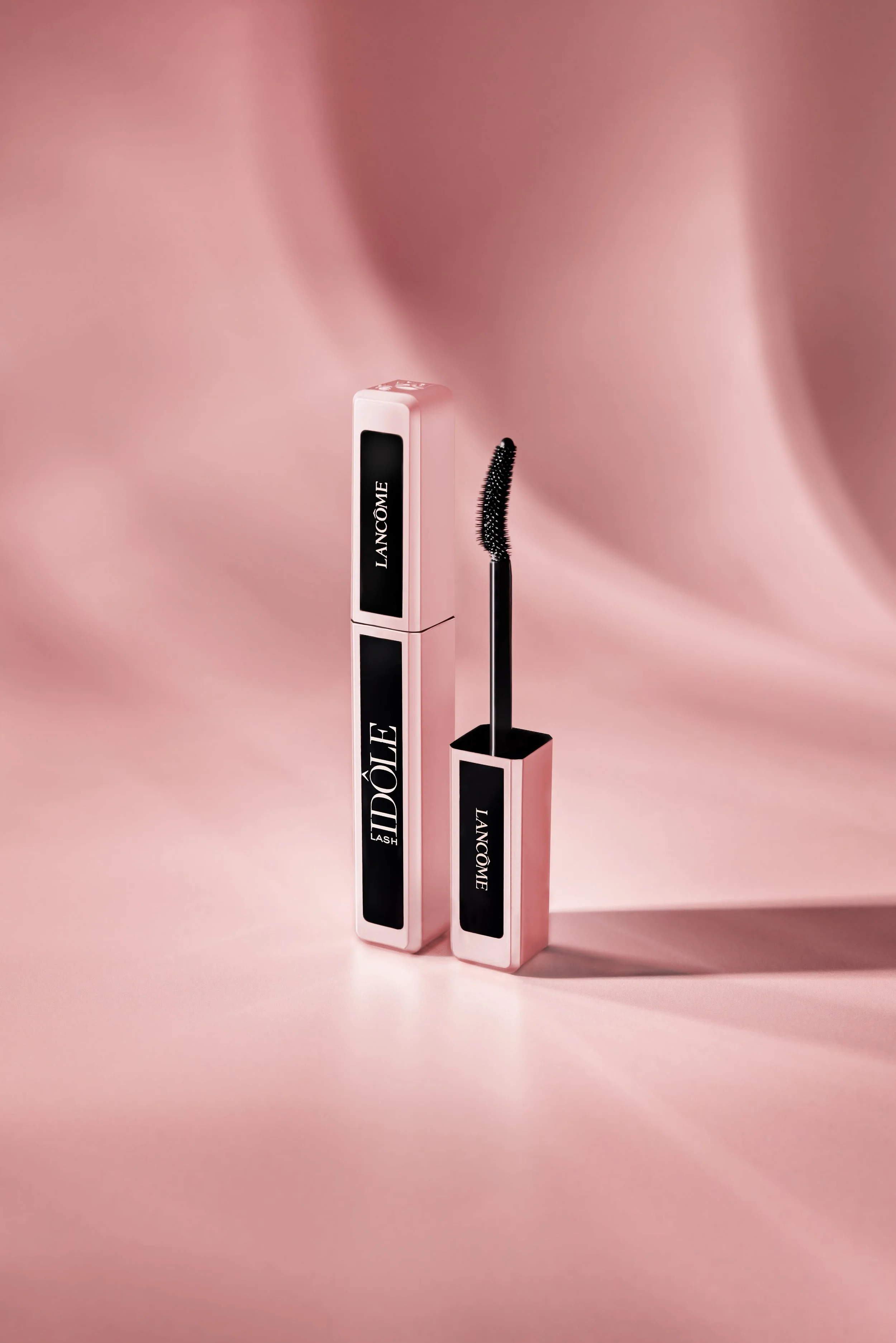 Lancôme Lash Idôle Mascara. The image features the sleek pink and black mascara tube standing upright alongside its curved mascara wand, set against a luxurious, draped silk background in matching rose tones.