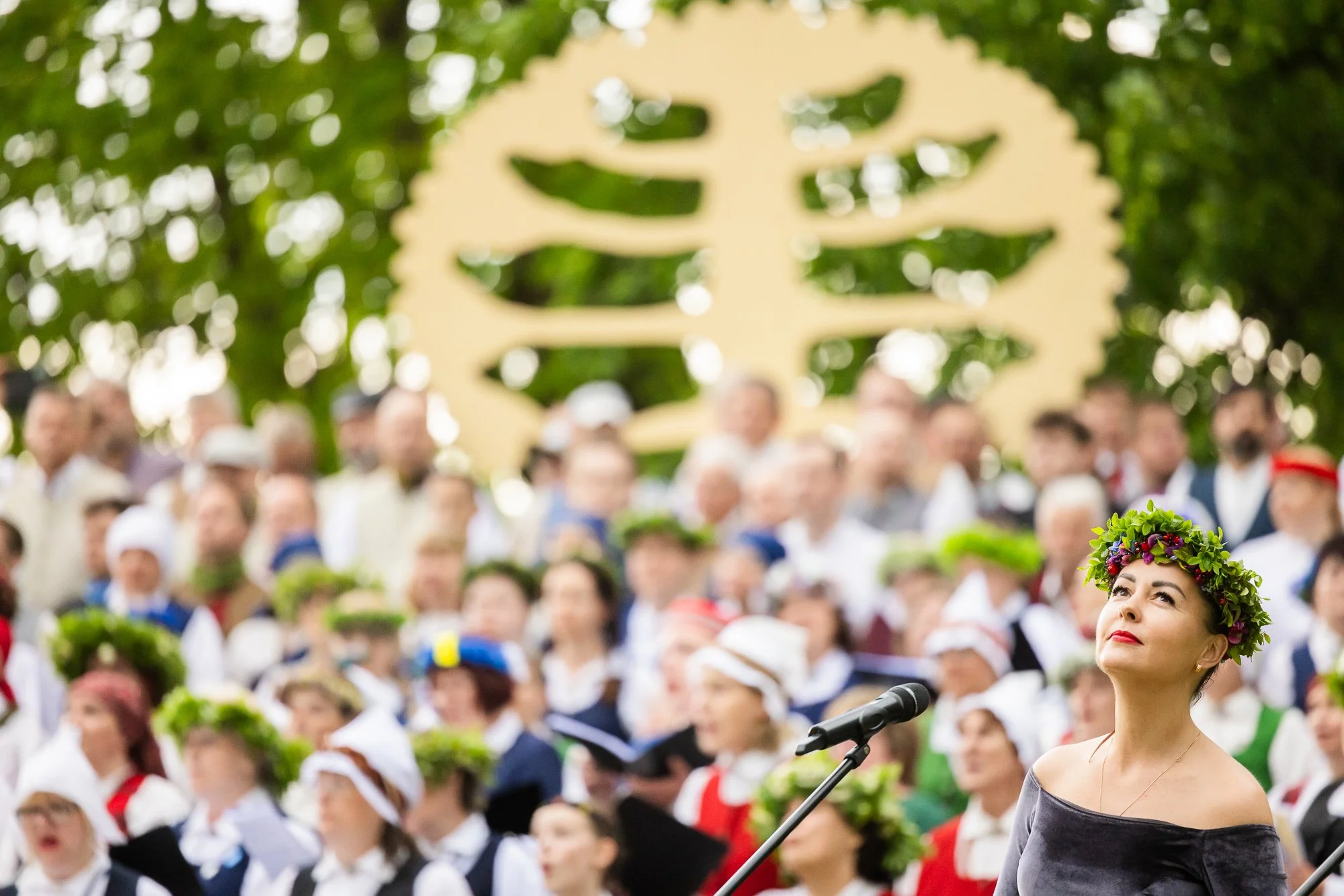 A woman wearing a flower crown stands in front of a large choir, speaking into a microphone during an outdoor event with trees and a wood sculpture in the background.