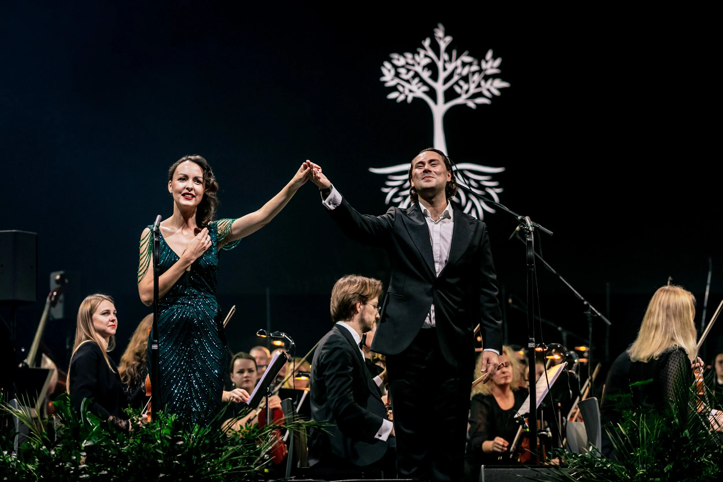 A man and woman holding hands on stage in front of an orchestra, with a white tree logo behind them.