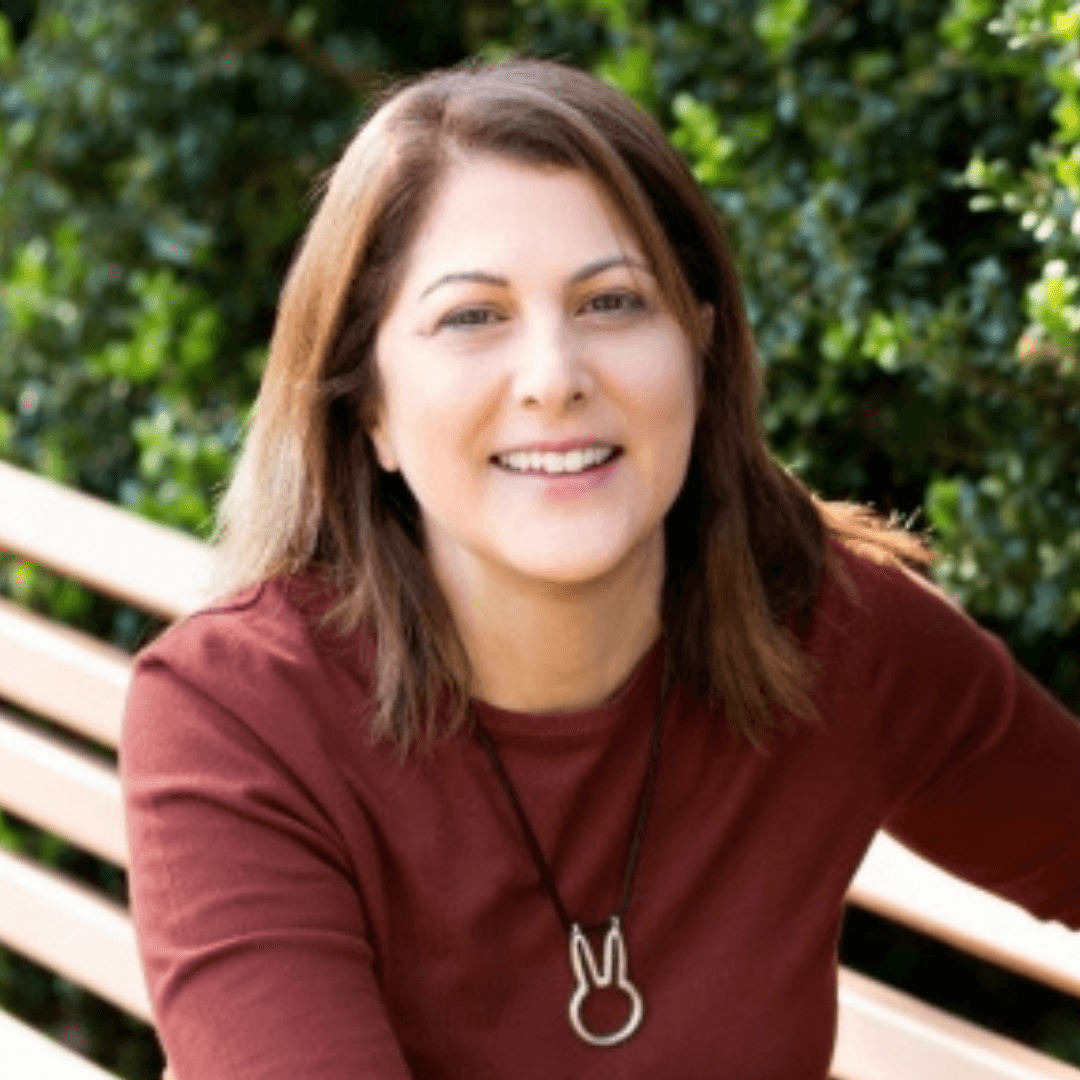 A woman with brown hair smiling, wearing a maroon top and a pendant necklace, sitting on a park bench with green foliage in the background.