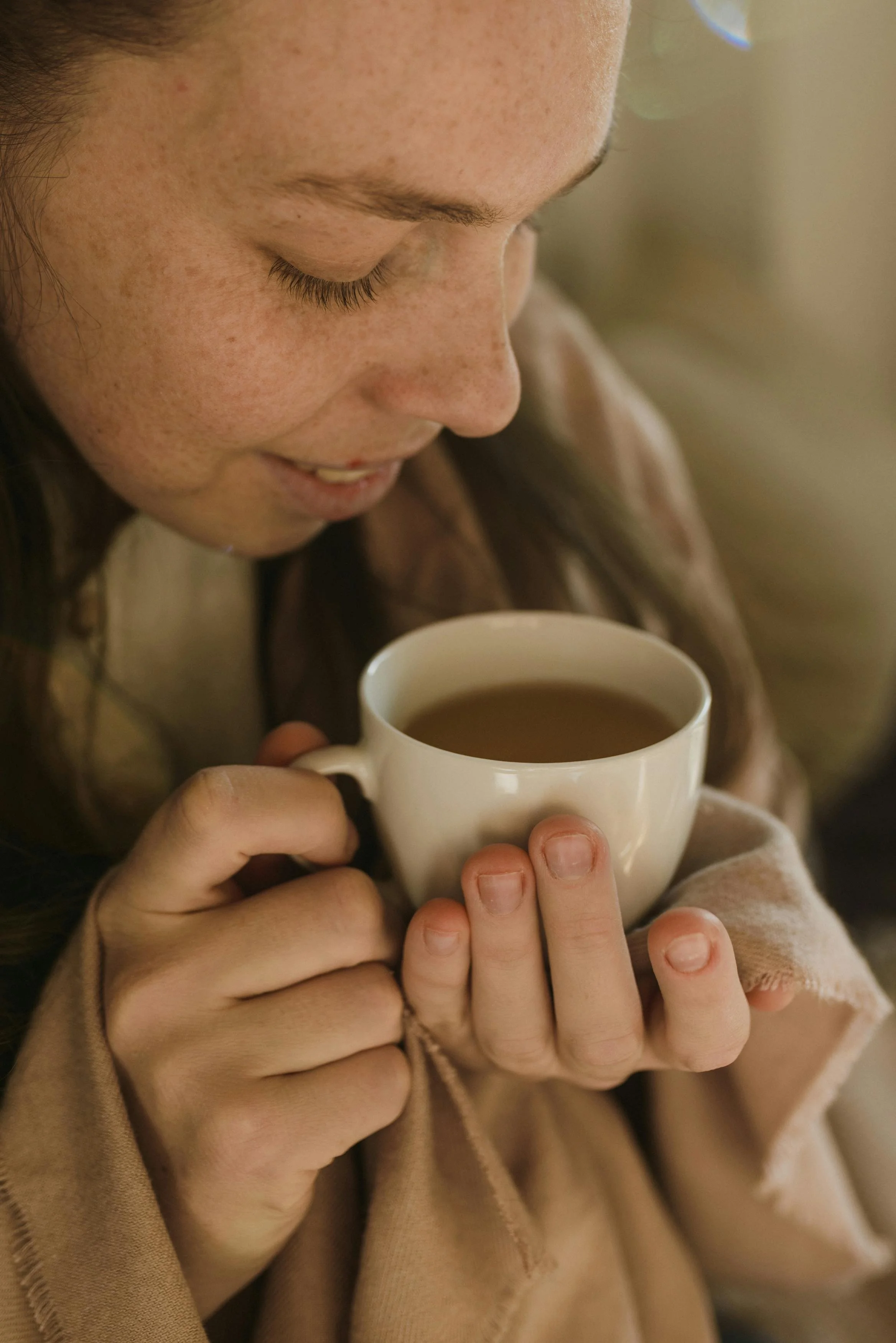 A woman with freckles holding a white mug of tea or coffee, smiling softly and looking down, dressed in warm clothing.