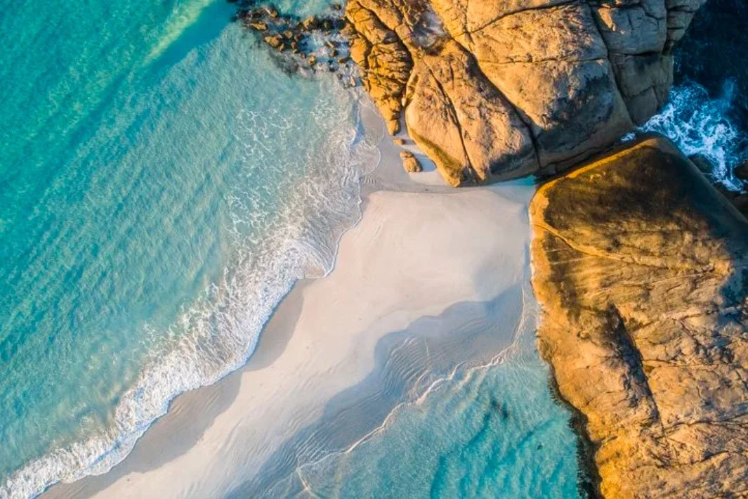 Aerial view of a beach with turquoise water, white sand, and large rocks along the shoreline at sunset.