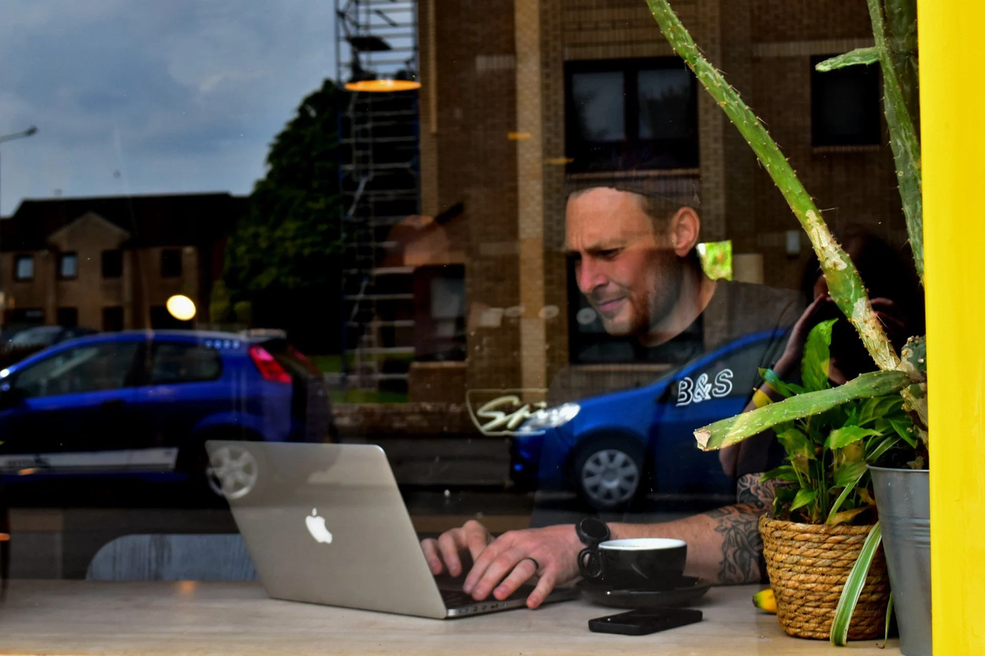 Greig photographed through the window of a coffee shop, drinking a coffee and working on remote running coaching services on his laptop
