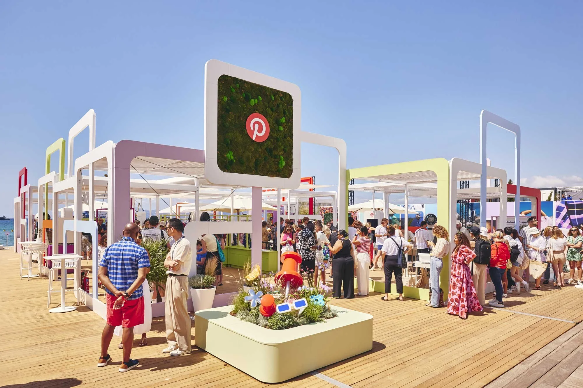 Crowd of people at an outdoor event on a wooden deck, with colorful modern structures and a Pinterest logo sign, under a clear blue sky.