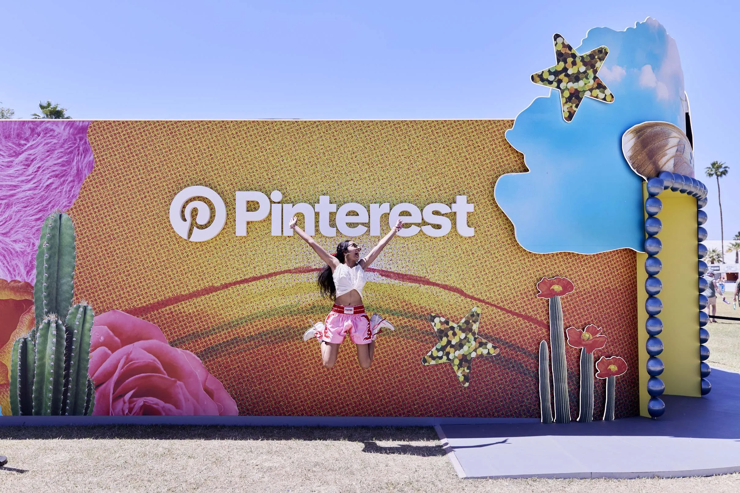 A woman jumping with arms raised in front of a Pinterest promotional display at an outdoor event. The display has large colorful decorations including cacti, stars, flowers, and a cloud with a star, with the Pinterest logo and text in the center.