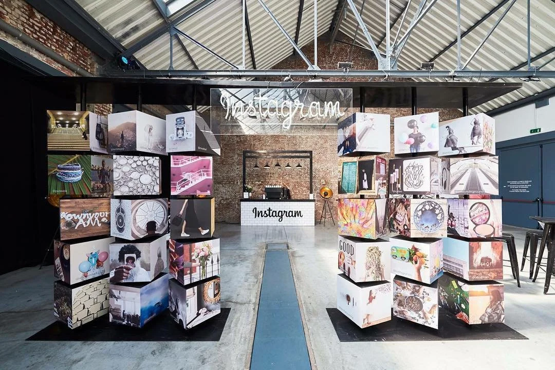 An indoor art installation with cubes displaying various photographs and artwork, framing an Instagram sign with neon lights. The space has a high ceiling with metal beams and a brick wall background.