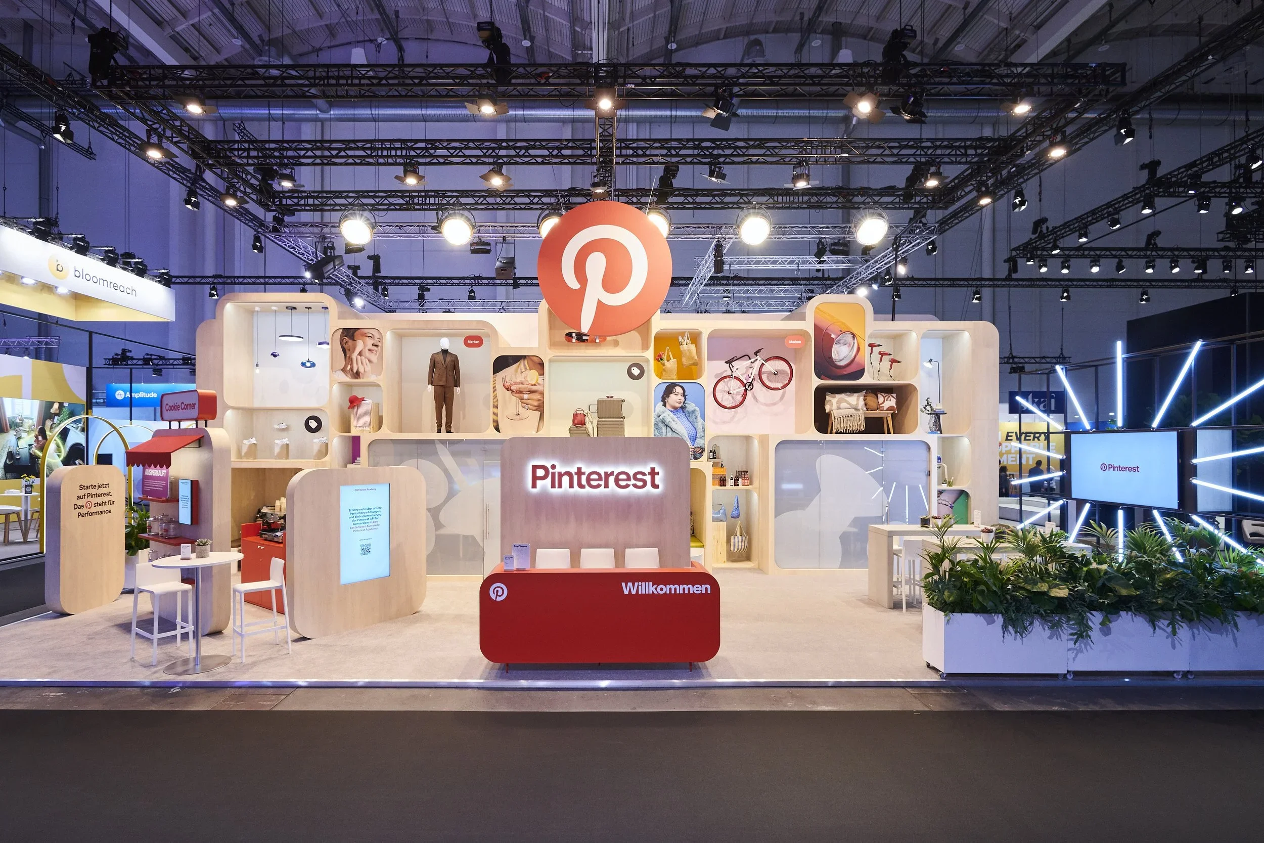 Exhibition booth for Pinterest featuring a large Pinterest logo, a welcome sign in German, and various products and displays, including a bicycle, a suit, and decorative items, under bright lighting at a trade show.
