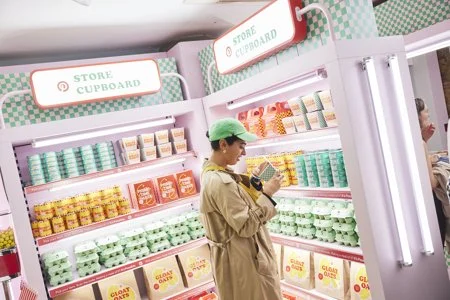A person wearing a green cap and tan coat shopping in a store with shelves of various colored containers, possibly yogurt or food items.