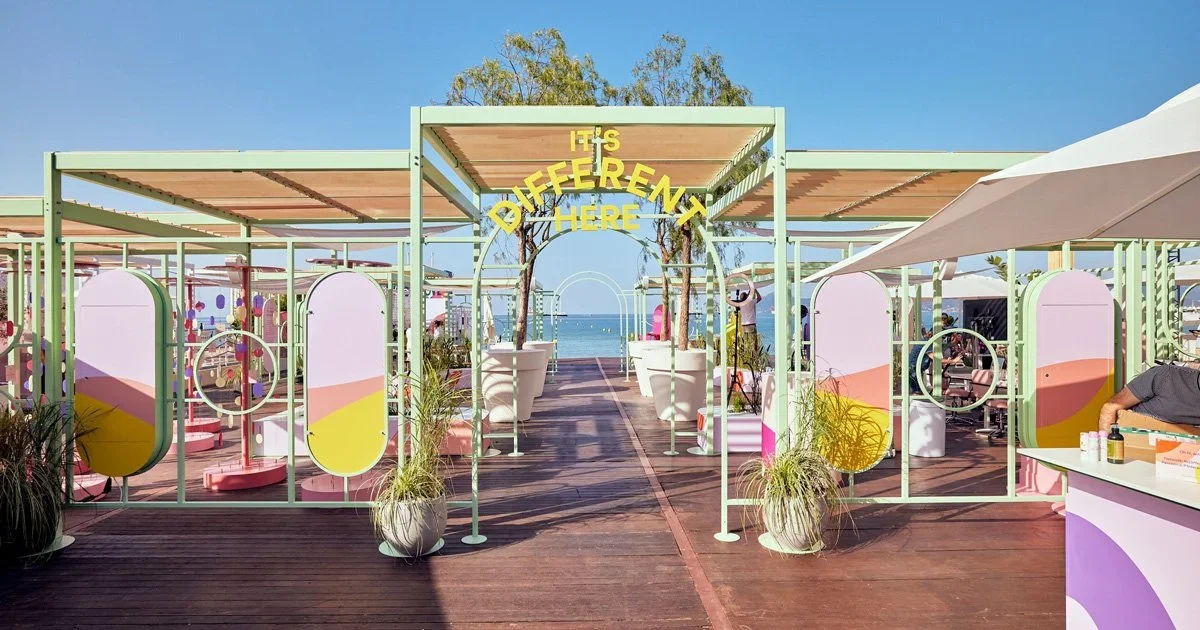 Colorful outdoor beachfront area with a sign reading 'It's Different Here', surrounded by pastel-colored structures, potted plants, and seaside view in the background.